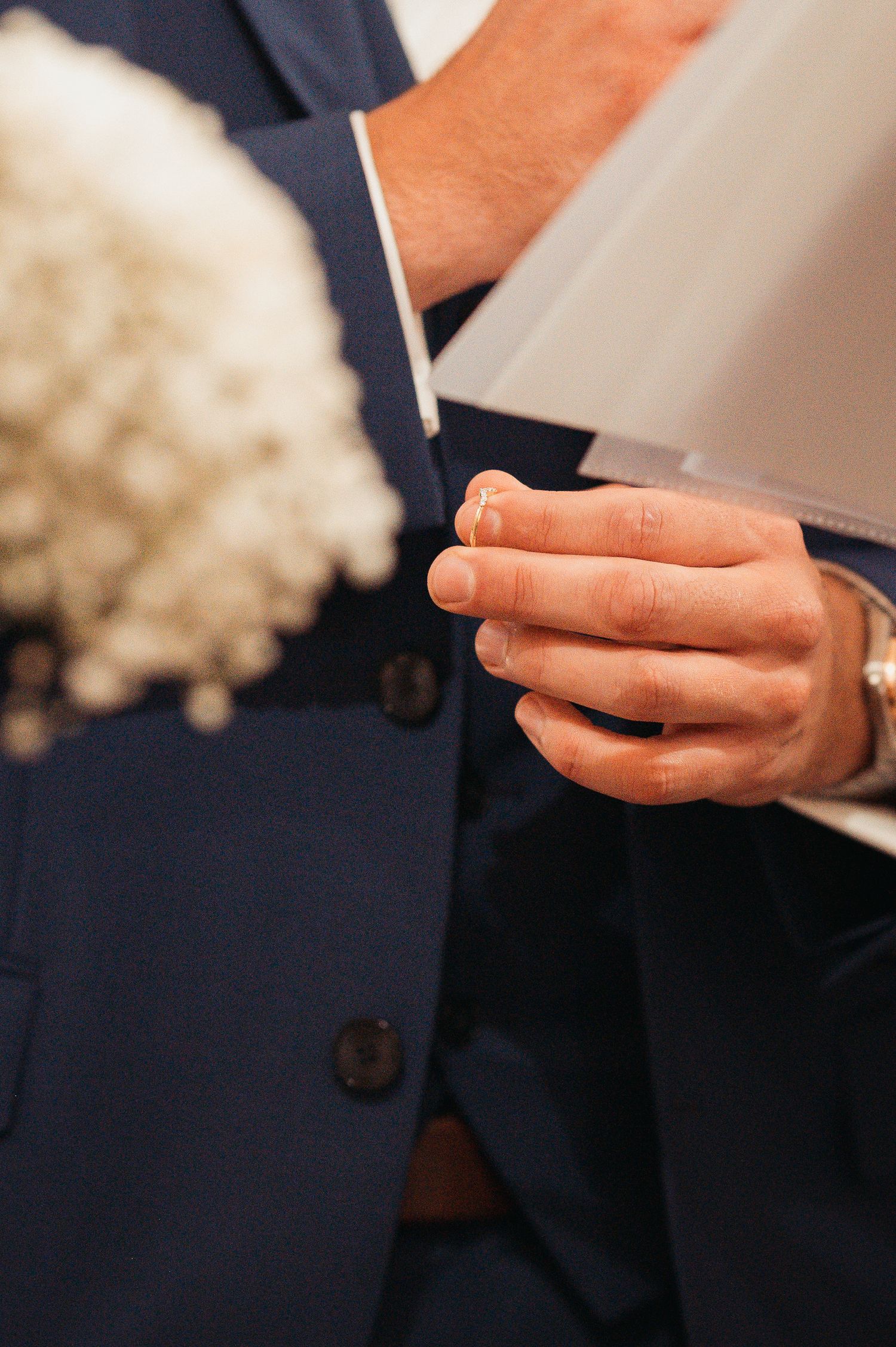 Groom in navy blue suit holding a wedding ring during ceremony, with white flowers visible in foreground.