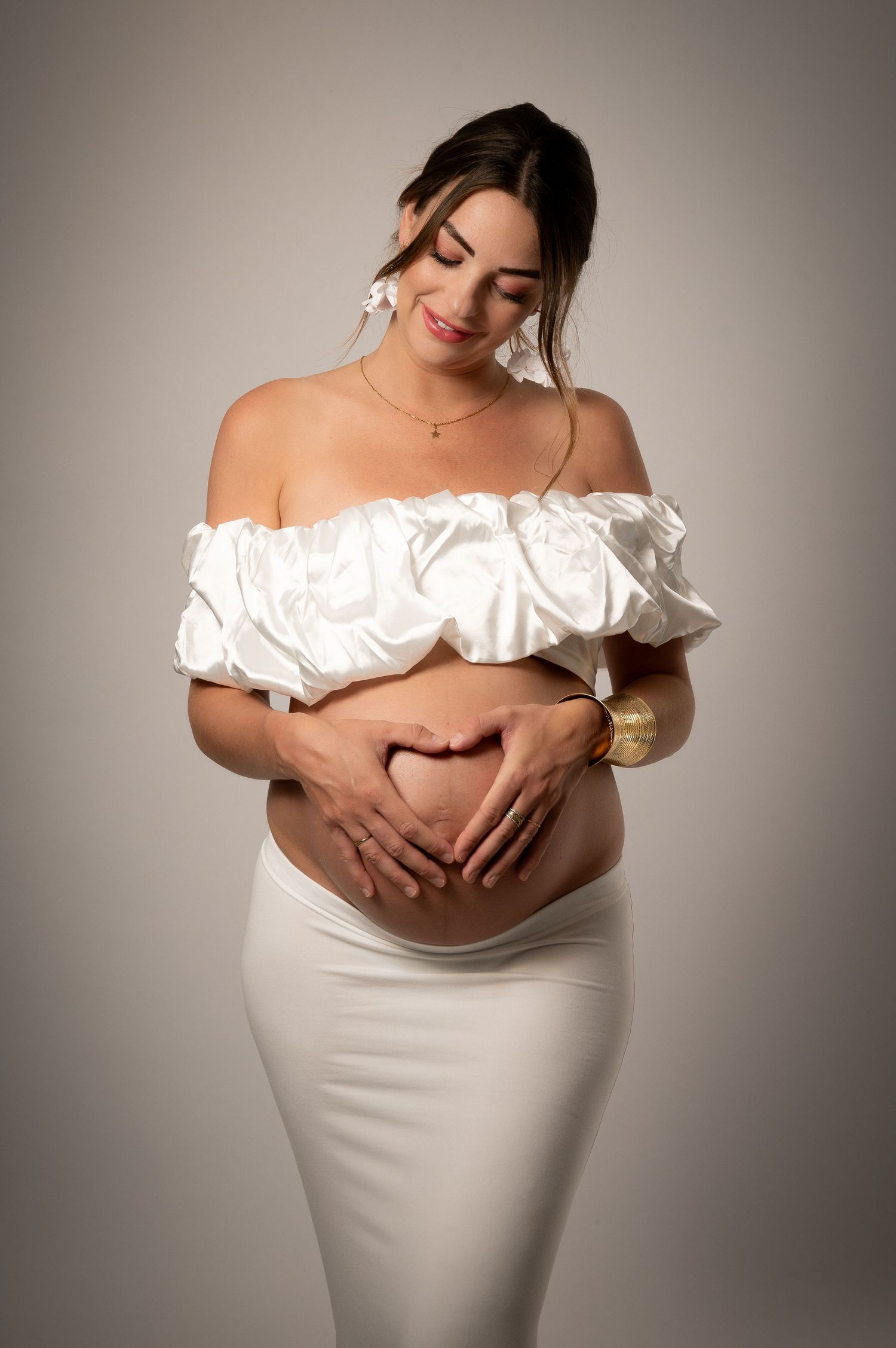 Pregnant woman in white off-shoulder top and skirt making heart shape with hands on baby bump, smiling softly.