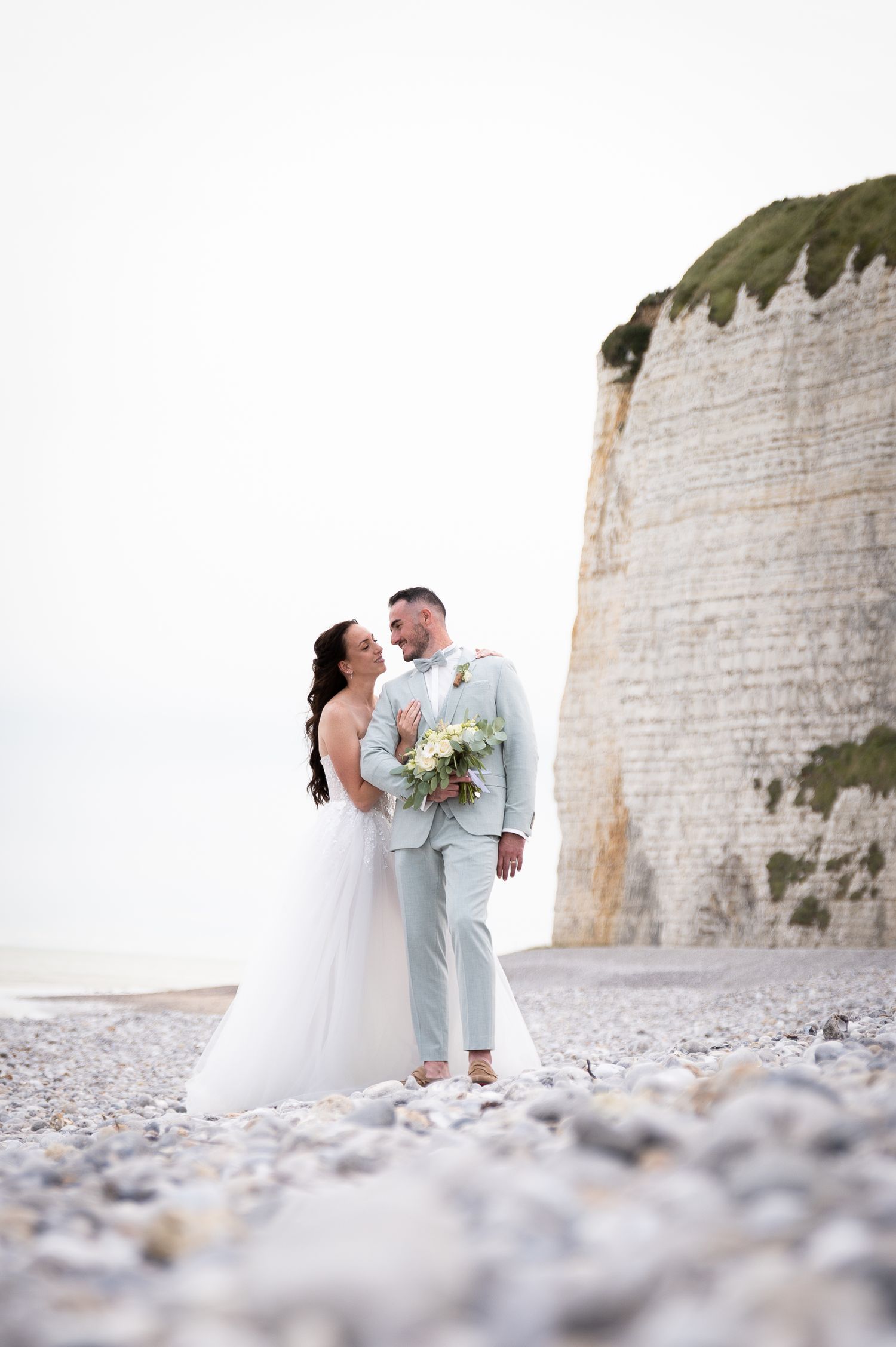 Bride and groom embrace on a pebble beach with white chalk cliffs, bride holding a bouquet in a white gown.
