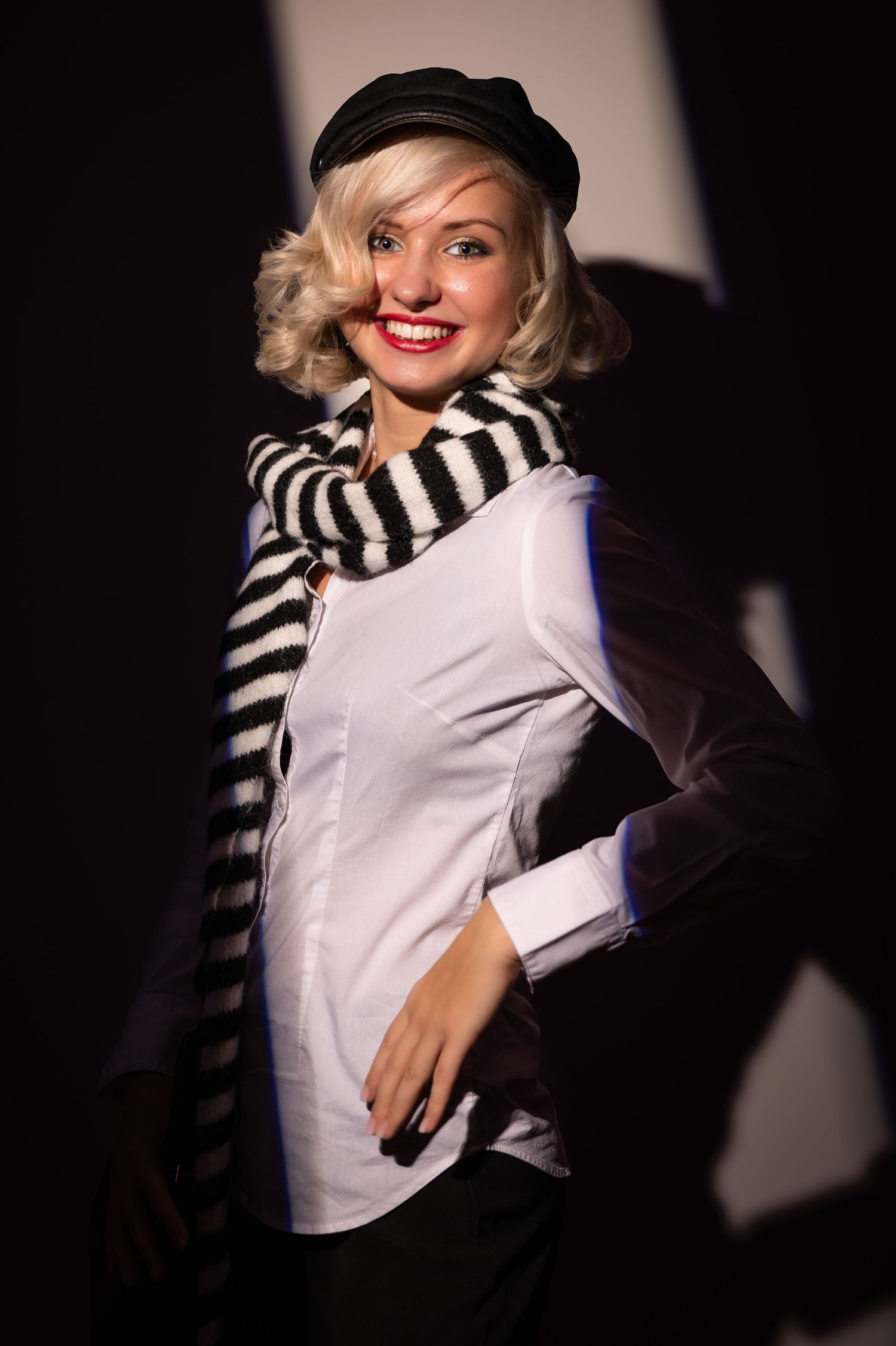 A smiling blonde woman in a black cap, striped scarf, and white shirt poses in dramatic shadow lighting.
