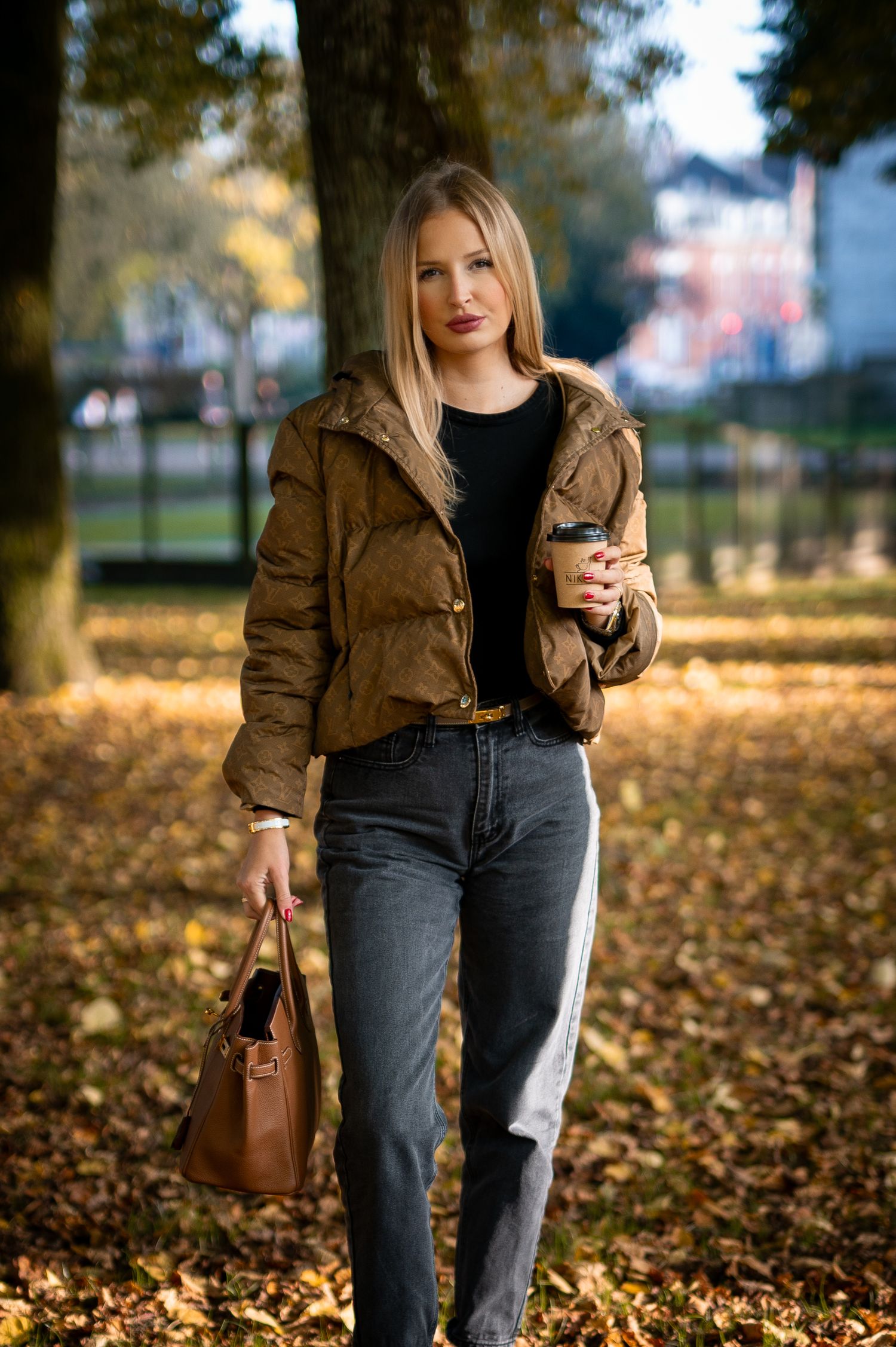 Young blonde woman in brown puffer jacket holding coffee cup and handbag walking in autumn park.