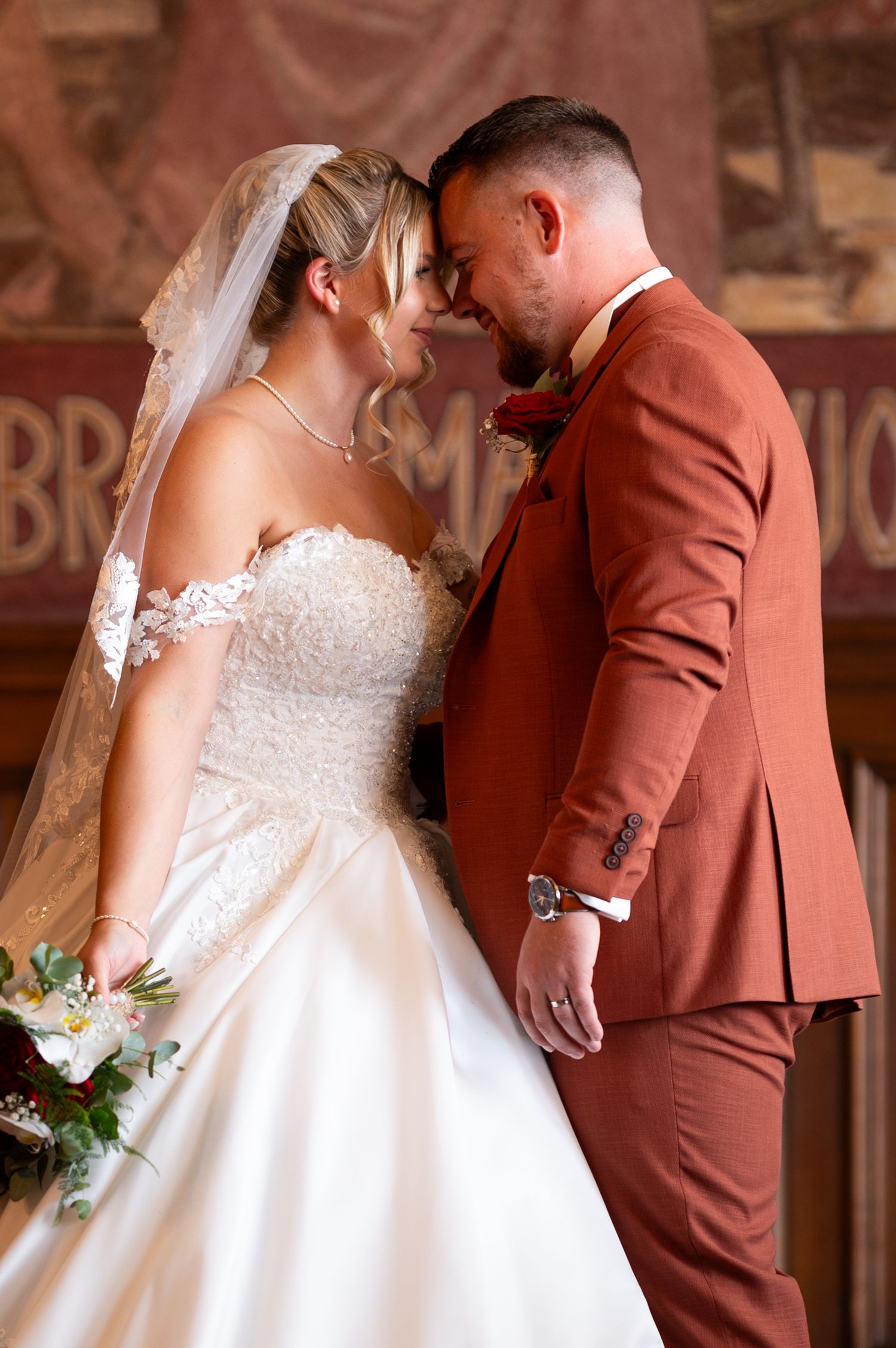 Bride in white lace gown and groom in rust suit touching foreheads tenderly, sharing romantic moment at wedding ceremony.