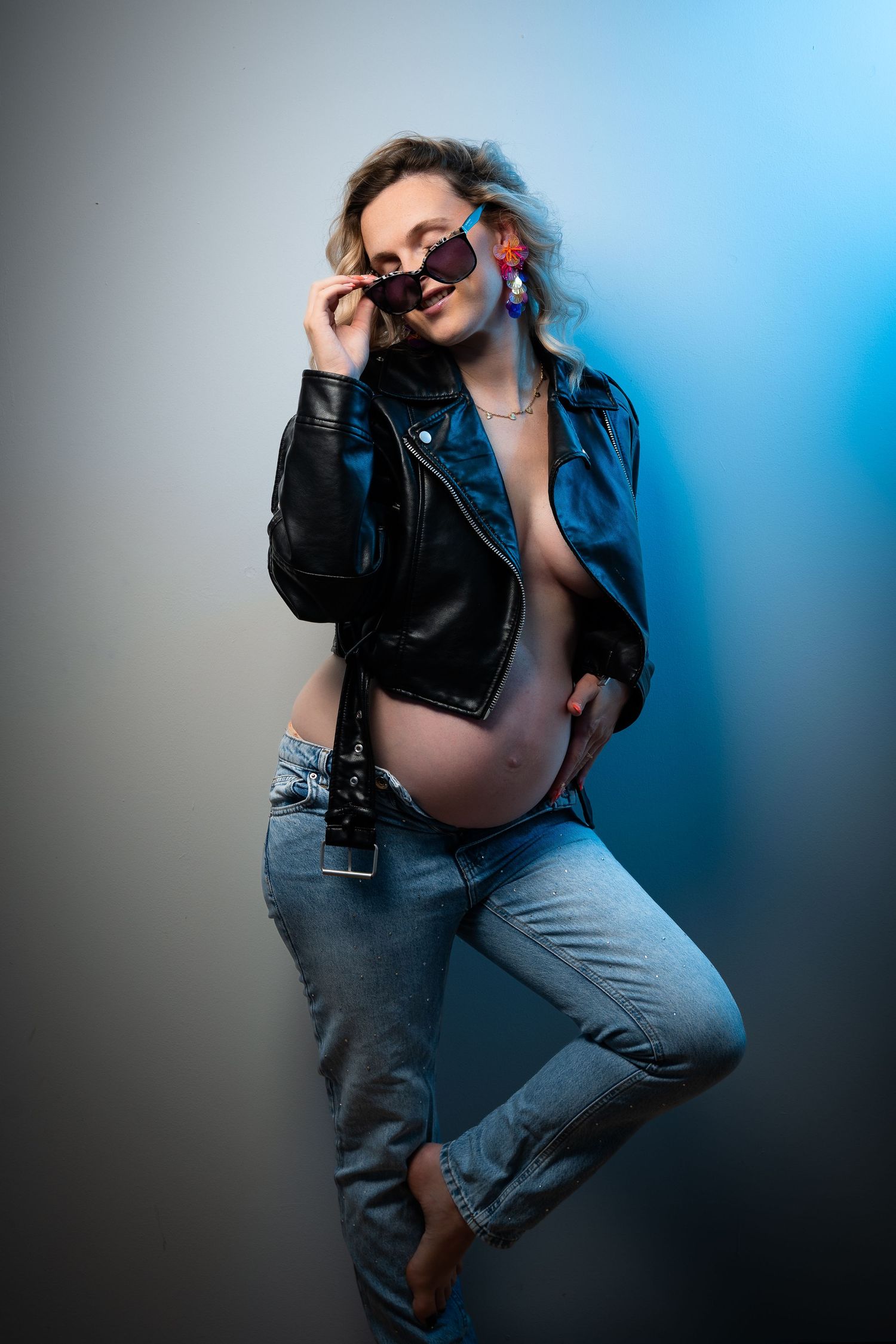 Pregnant woman in leather jacket, jeans, and sunglasses posing confidently in a studio with blue lighting.