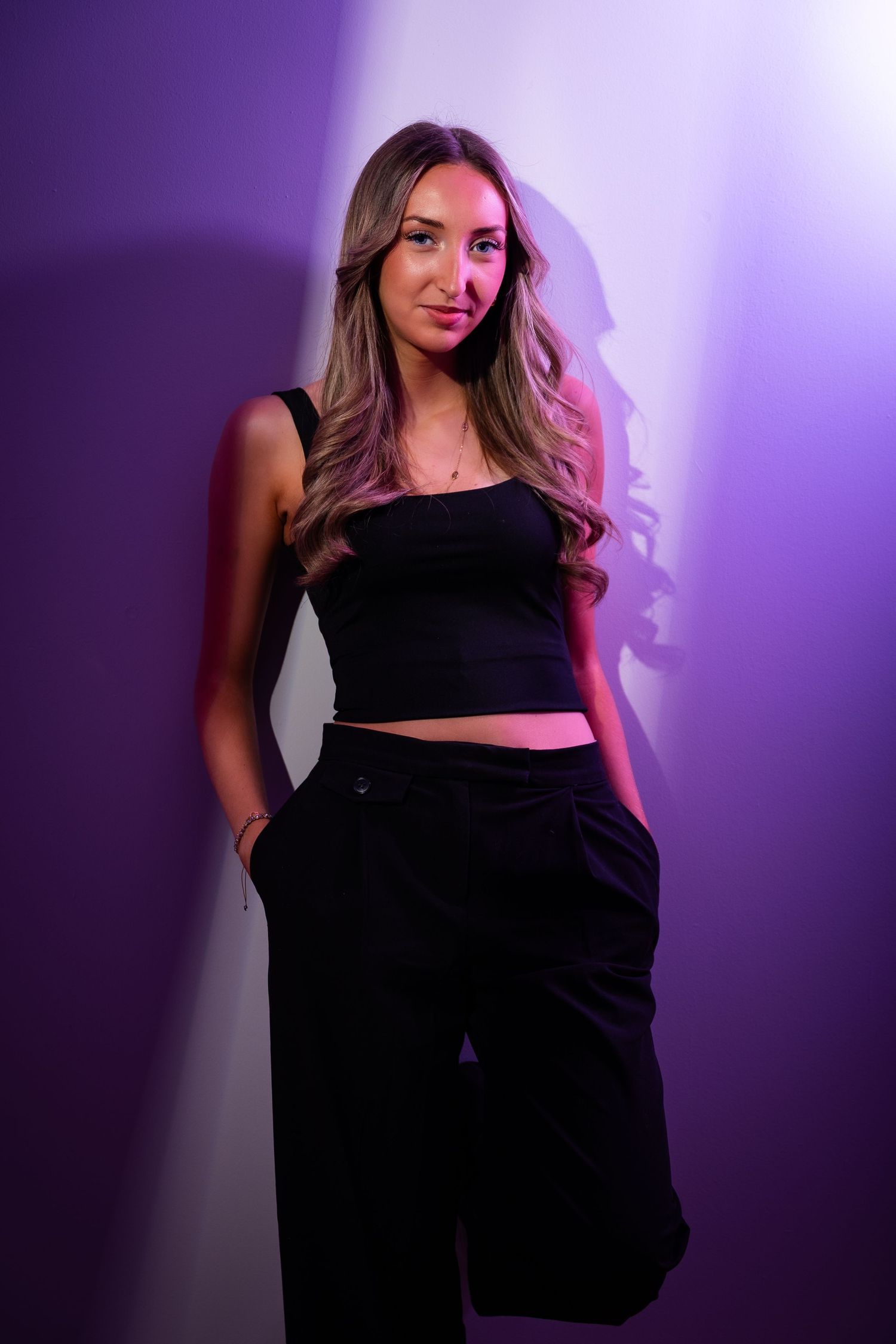 Young woman in black crop top and wide-leg pants poses confidently against a purple-lit studio wall.