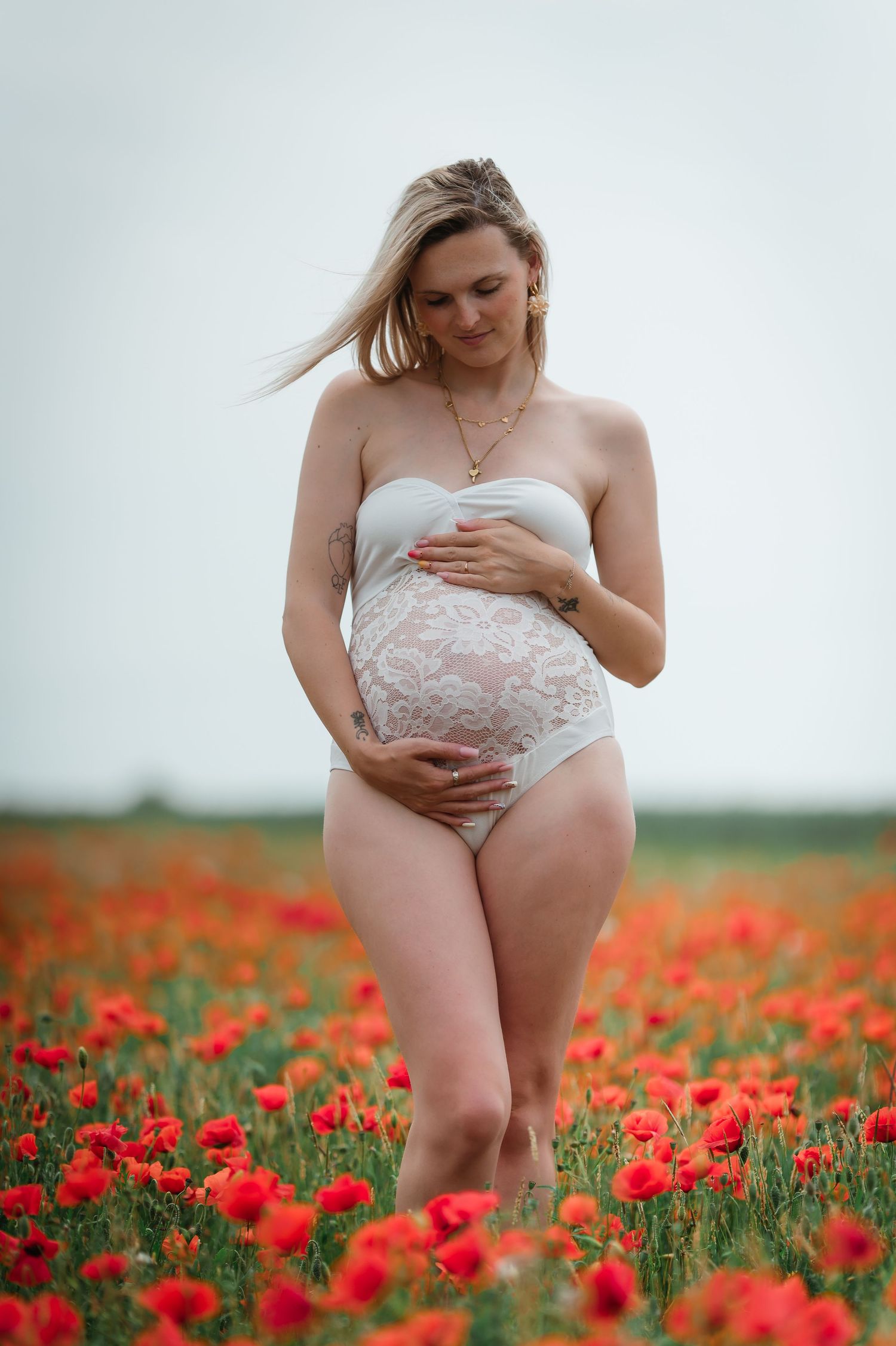 Pregnant woman in white lace lingerie standing in a vibrant red poppy field, cradling her baby bump.