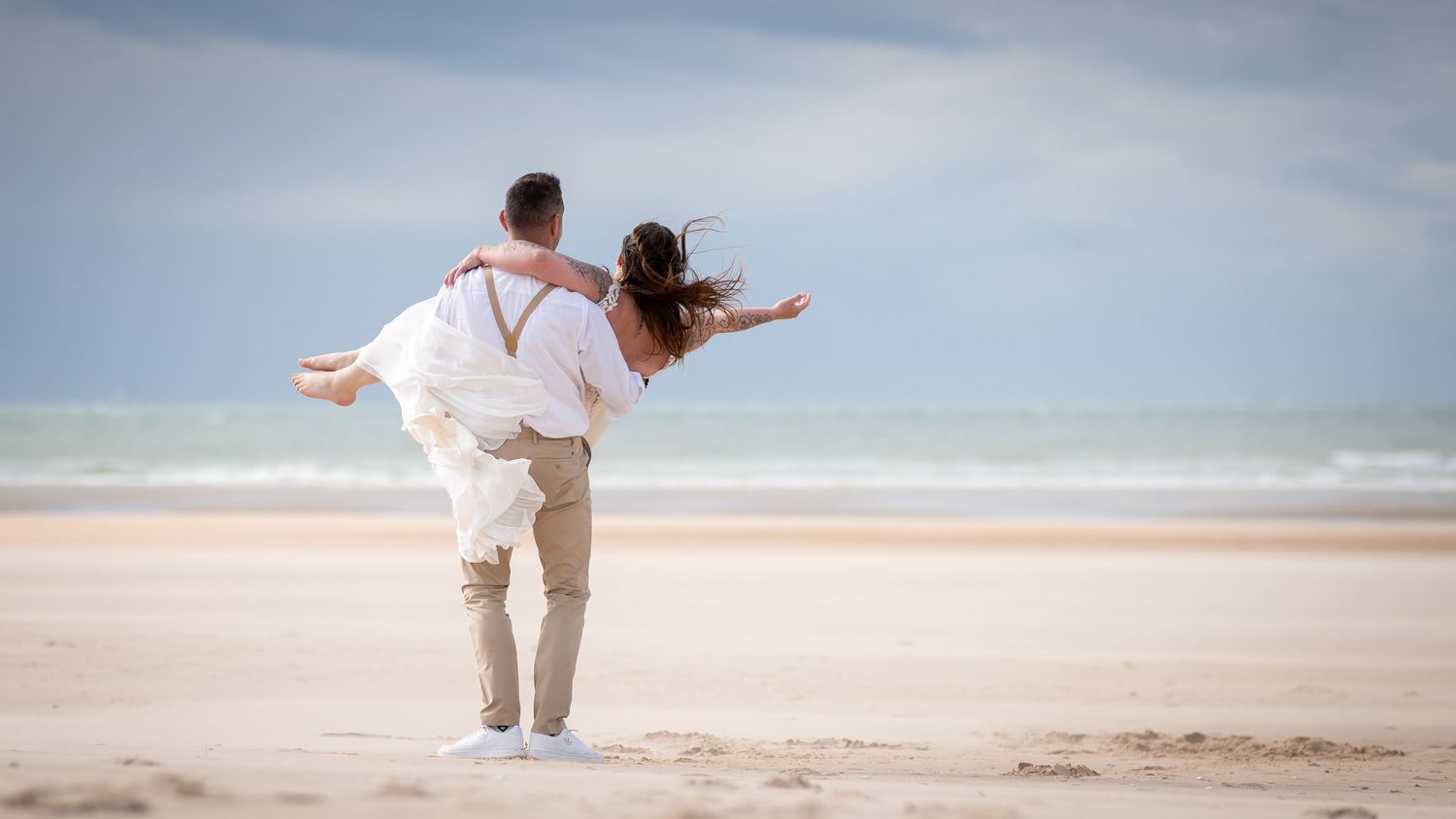 A couple in white wedding attire embrace joyfully on a wide sandy beach under a cloudy sky.
