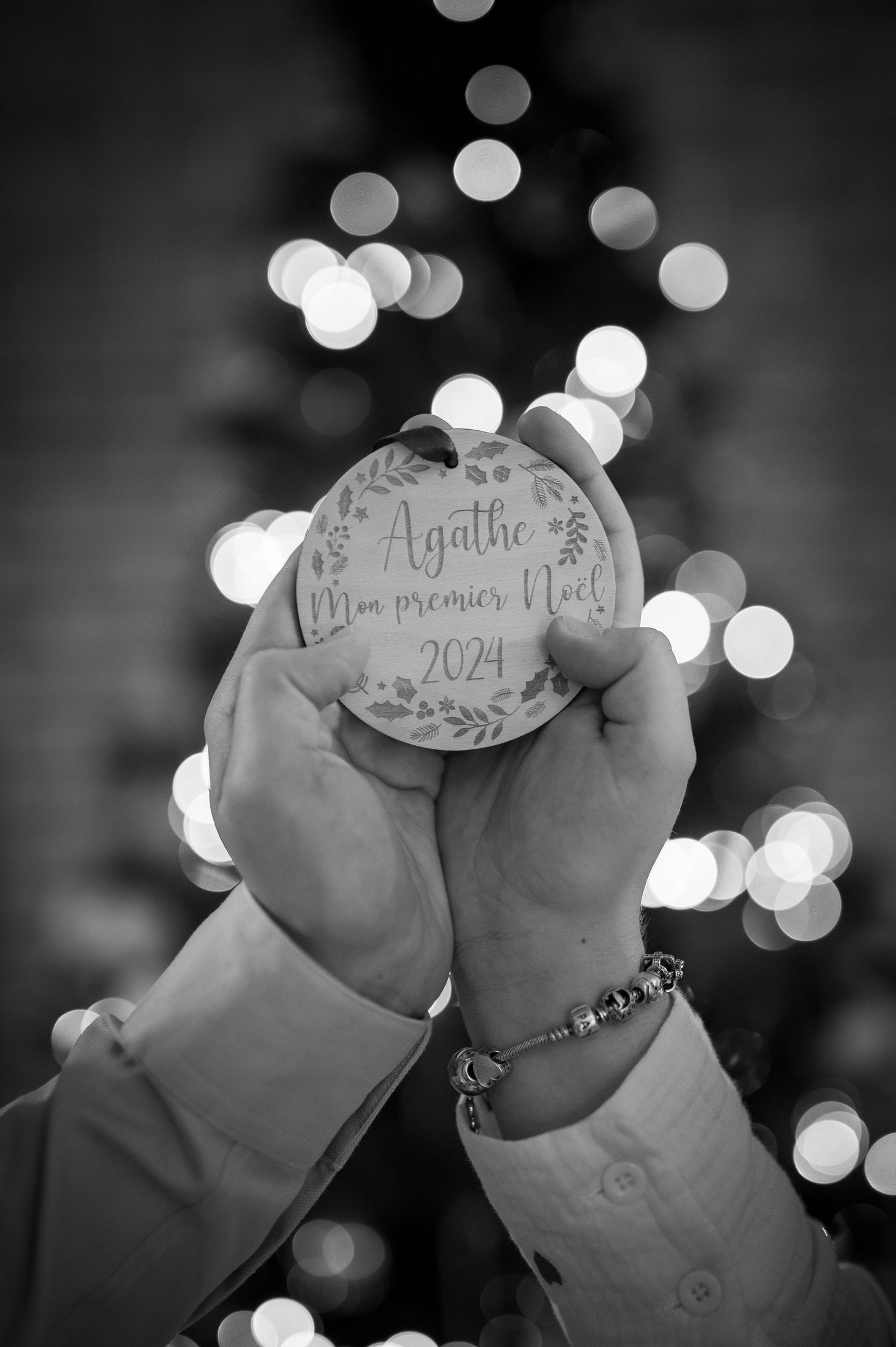 Black and white photo of hands holding a personalized Christmas ornament reading Agathe Mon premier Noel 2024 with bokeh lights.