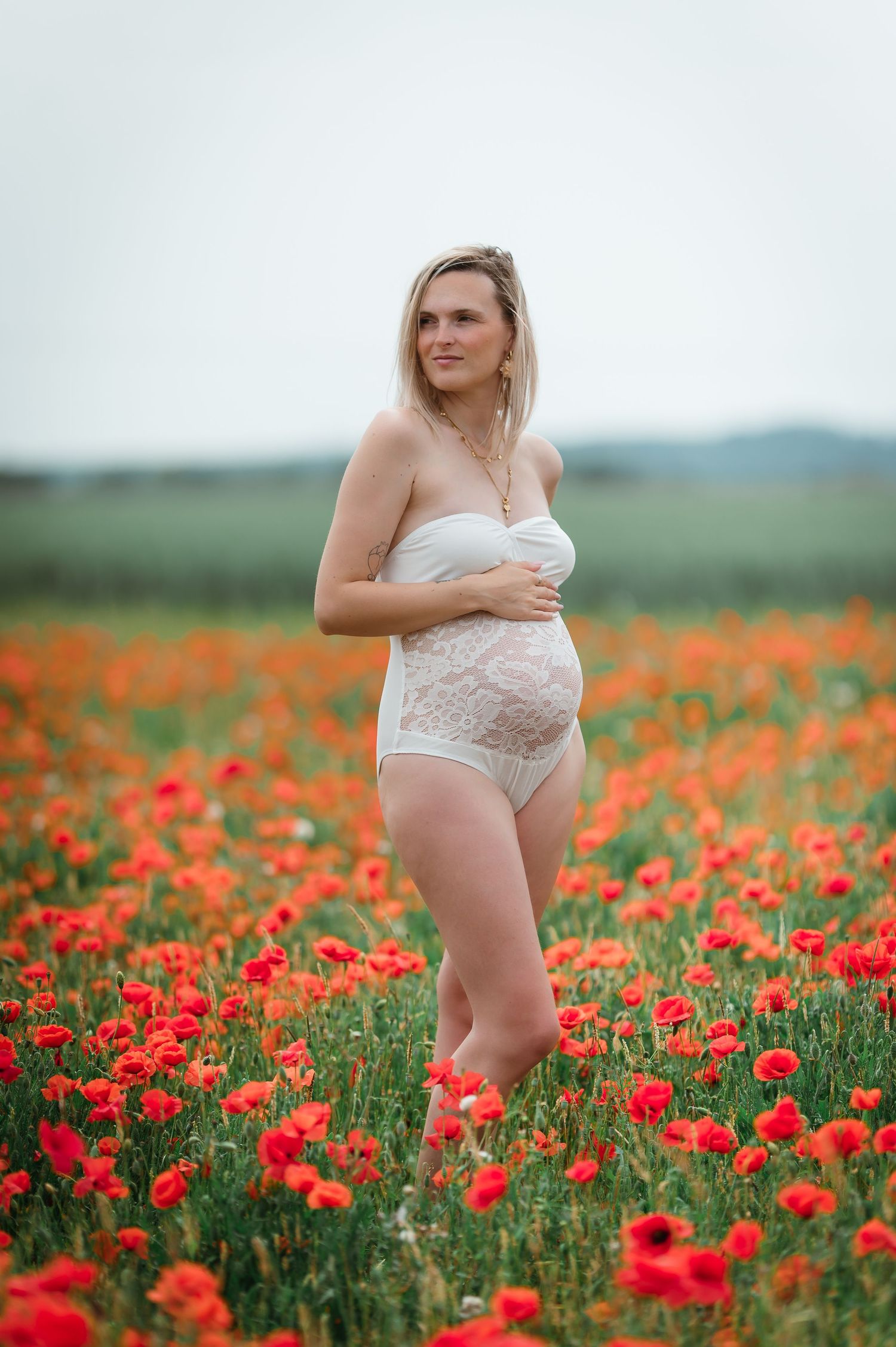 Pregnant woman in white lace lingerie standing in a vibrant red poppy field outdoors.