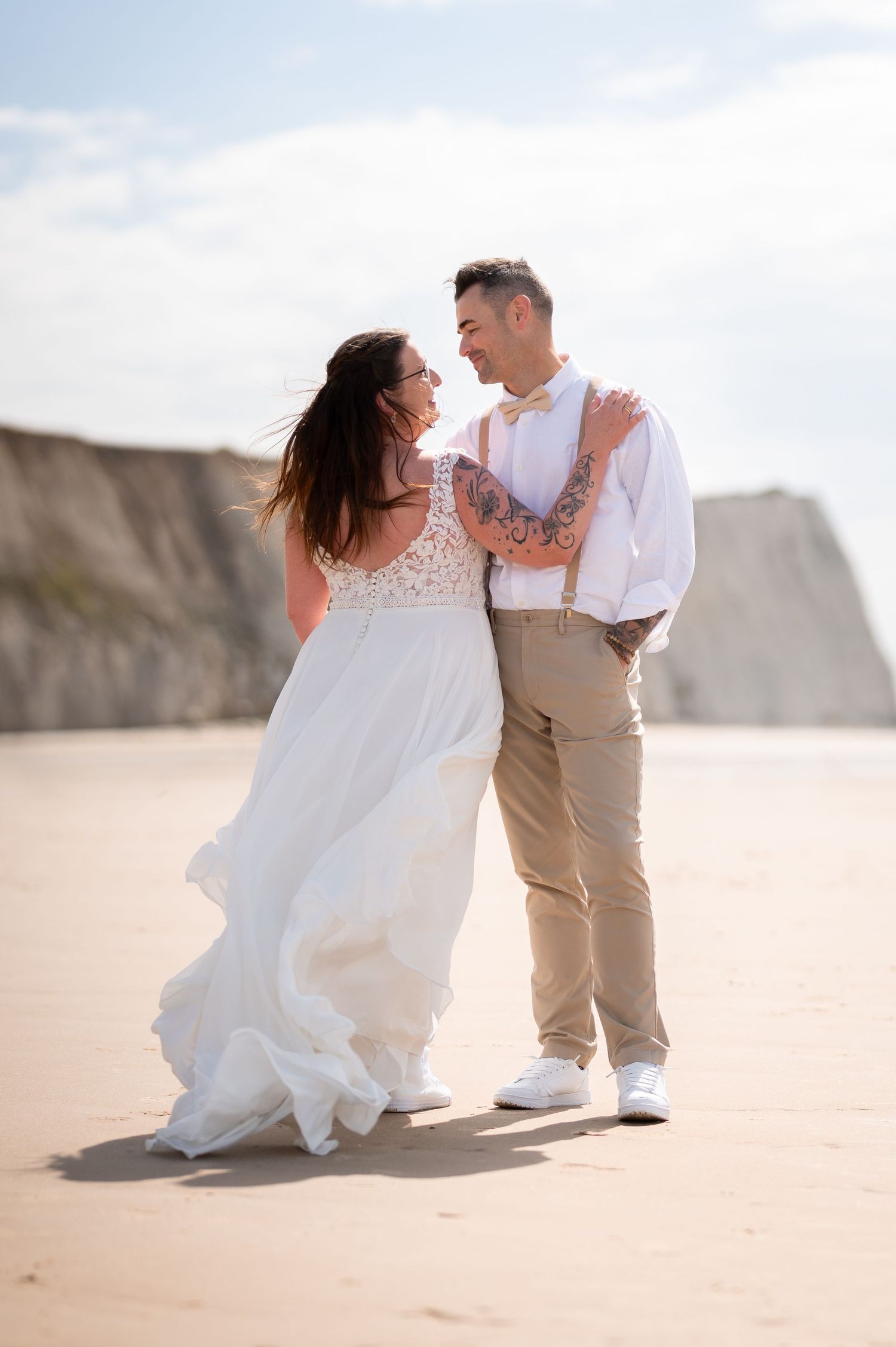 Bride in elegant white dress and groom share a tender moment on a sandy beach.