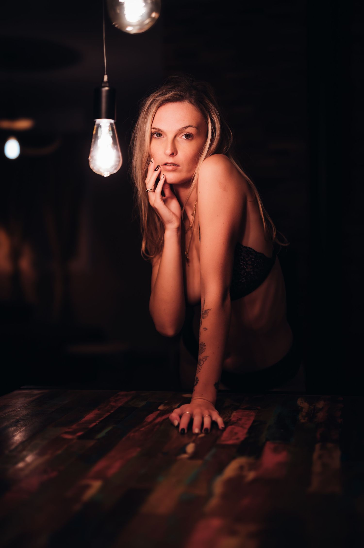 A blonde woman leans on a rustic wooden table under a glowing Edison bulb in a dark, moody setting.