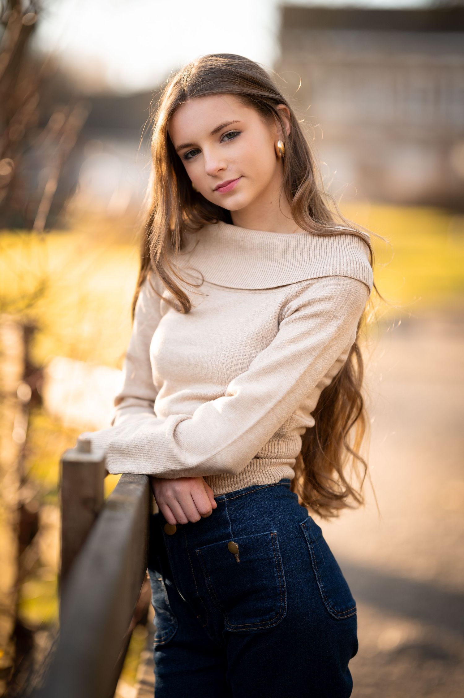 Young woman with long hair wearing a cream off-shoulder sweater and jeans, posing outdoors in warm sunlight.