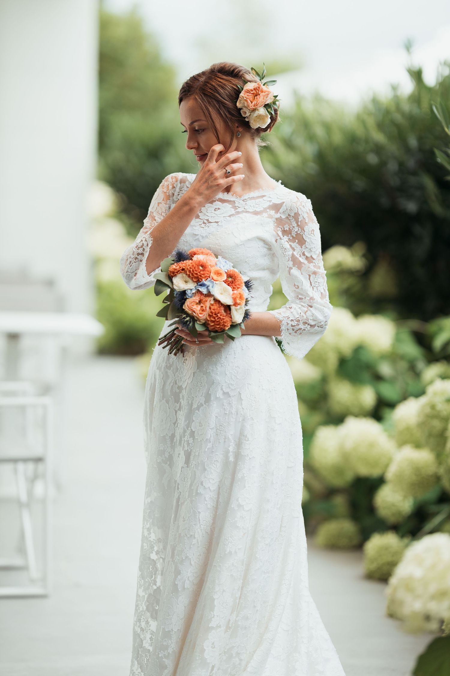 Bride in white lace gown holding orange bouquet, standing outdoors among hydrangeas with flower in her hair.