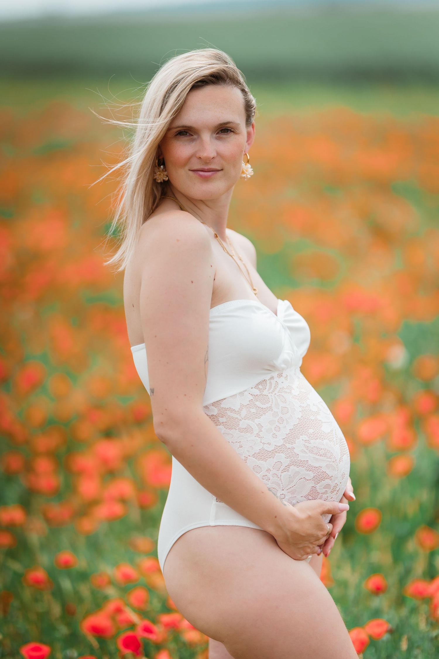 Pregnant woman in white lace bodysuit cradling baby bump in vibrant red poppy field.