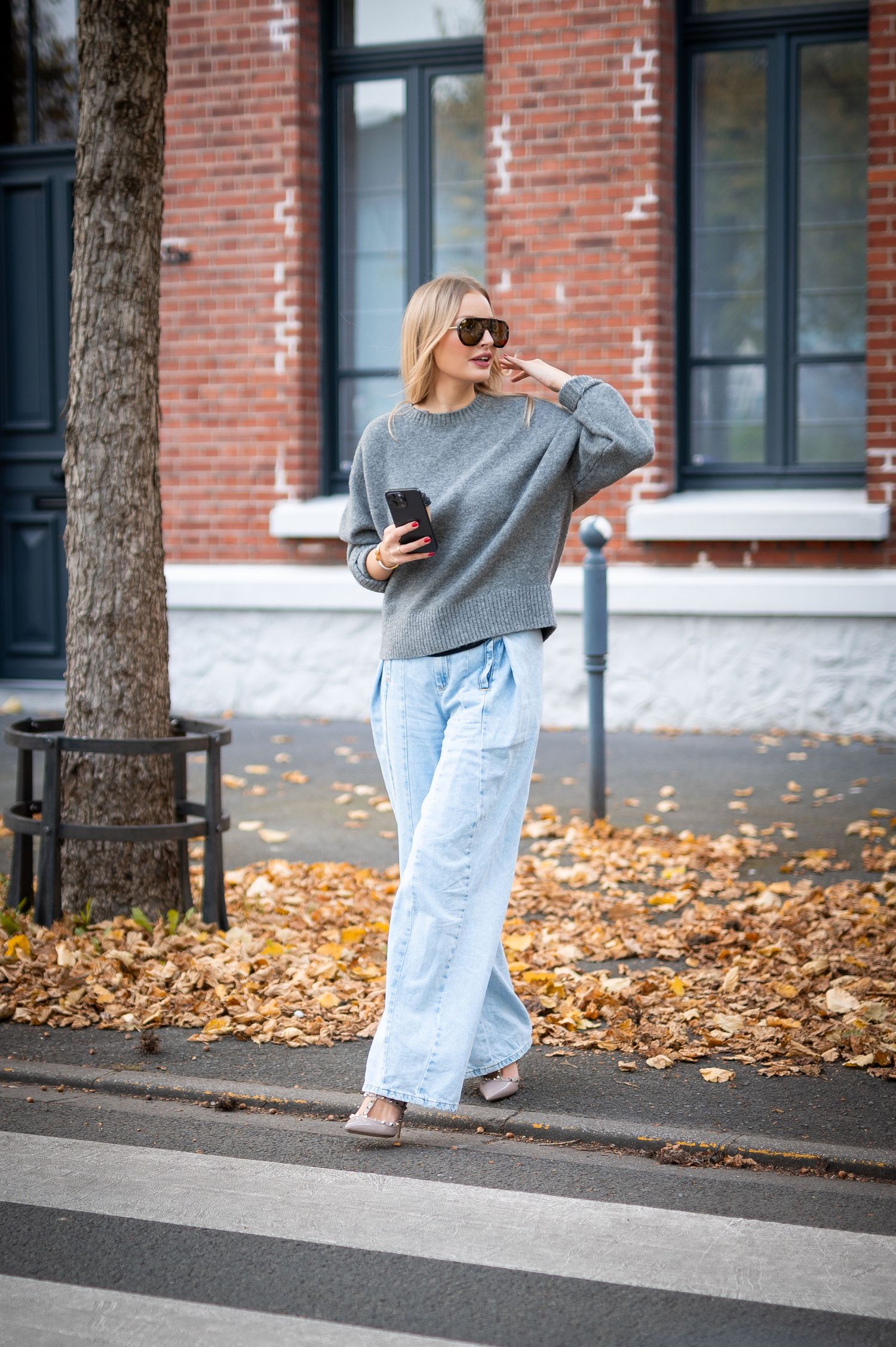 Blonde woman in gray sweater and wide-leg jeans crossing street in autumn, holding phone, wearing sunglasses.