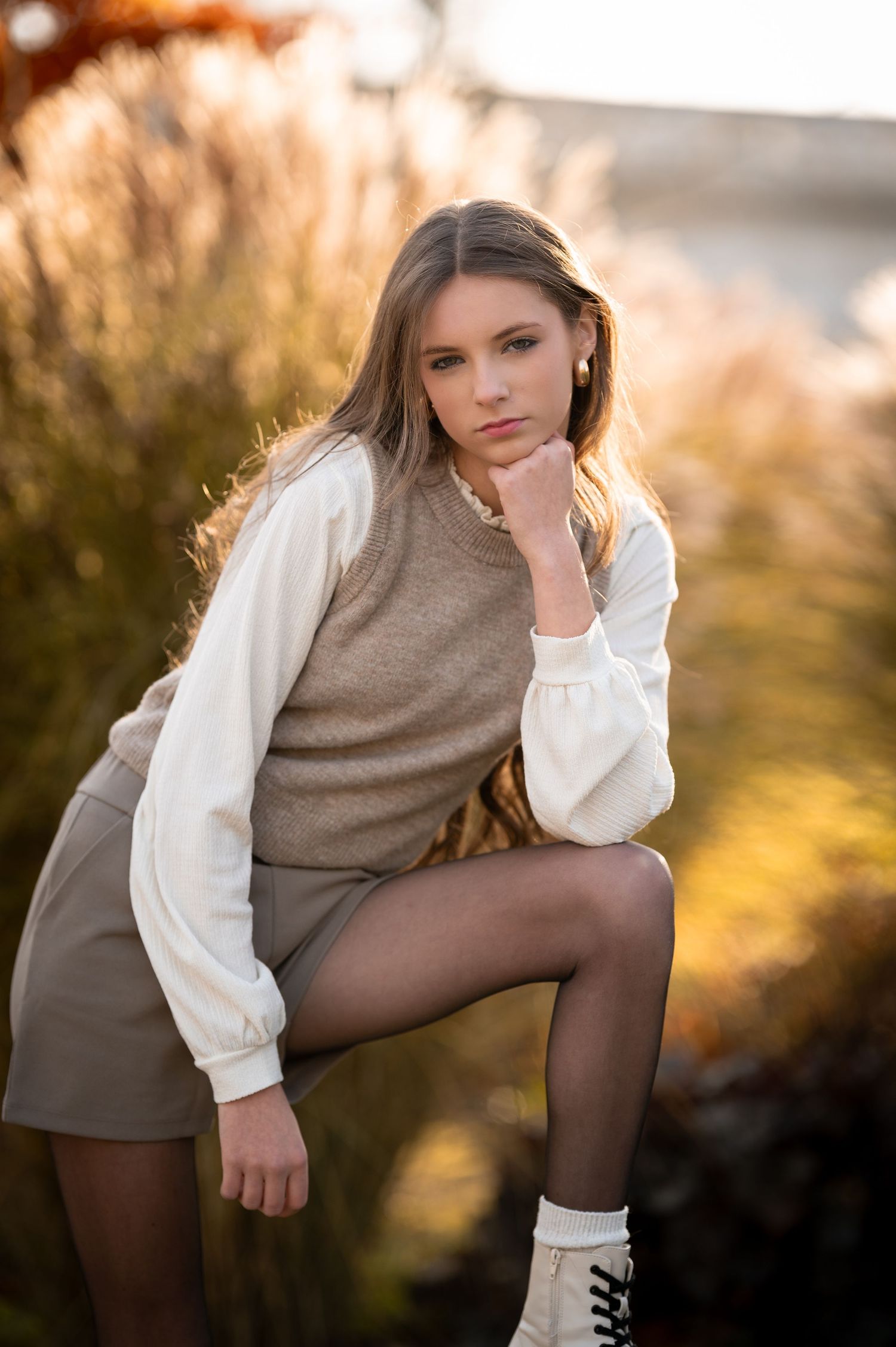 Young woman in tan sweater vest, white blouse, mini skirt, and boots poses outdoors in warm golden light.
