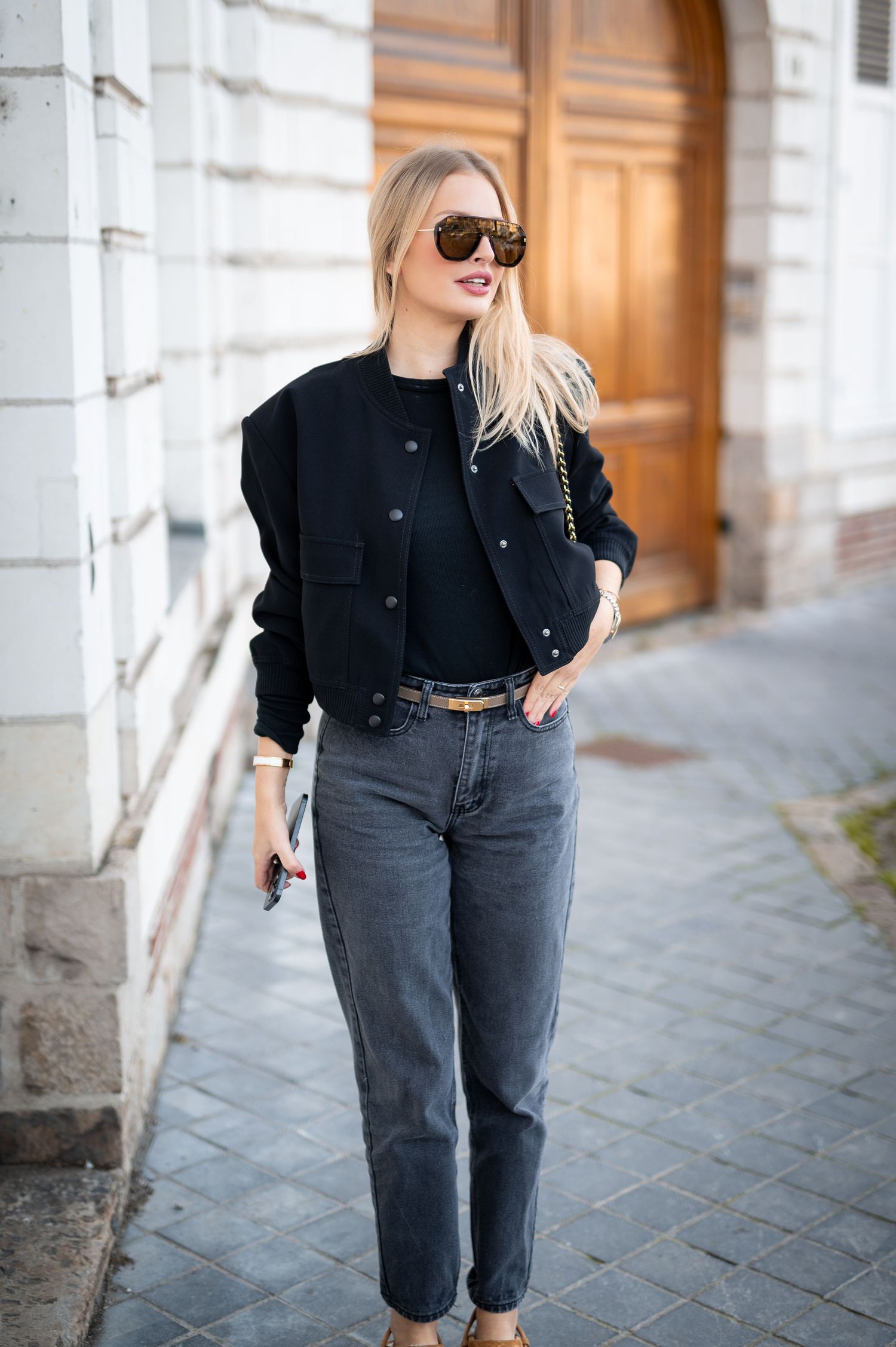 Blonde woman in black bomber jacket, dark jeans, and sunglasses posing on a cobblestone street.