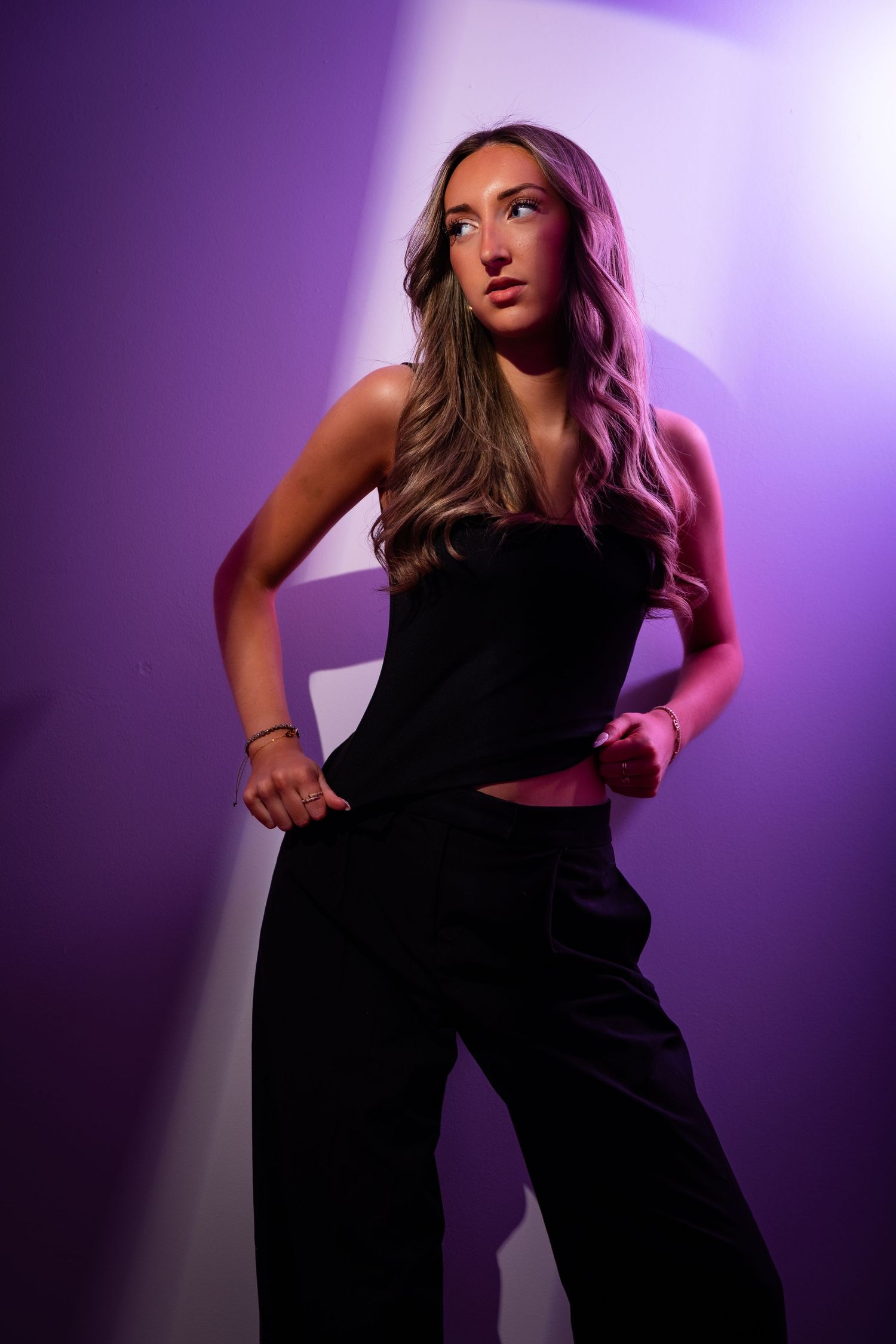 Young woman in black strapless top and wide-leg pants posing confidently under purple studio lighting.