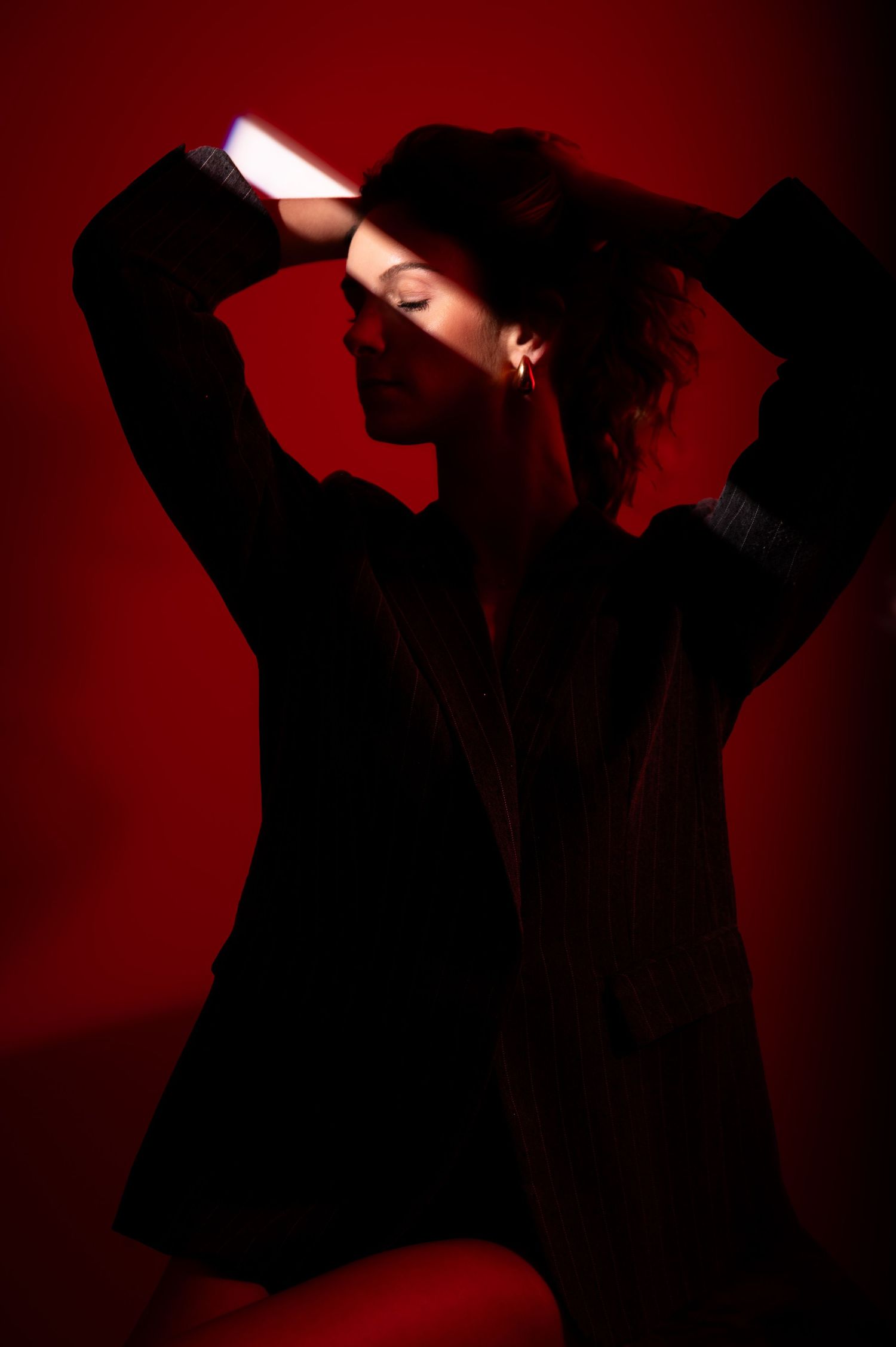 Woman in black blazer poses dramatically against red background with glowing light bar overhead.