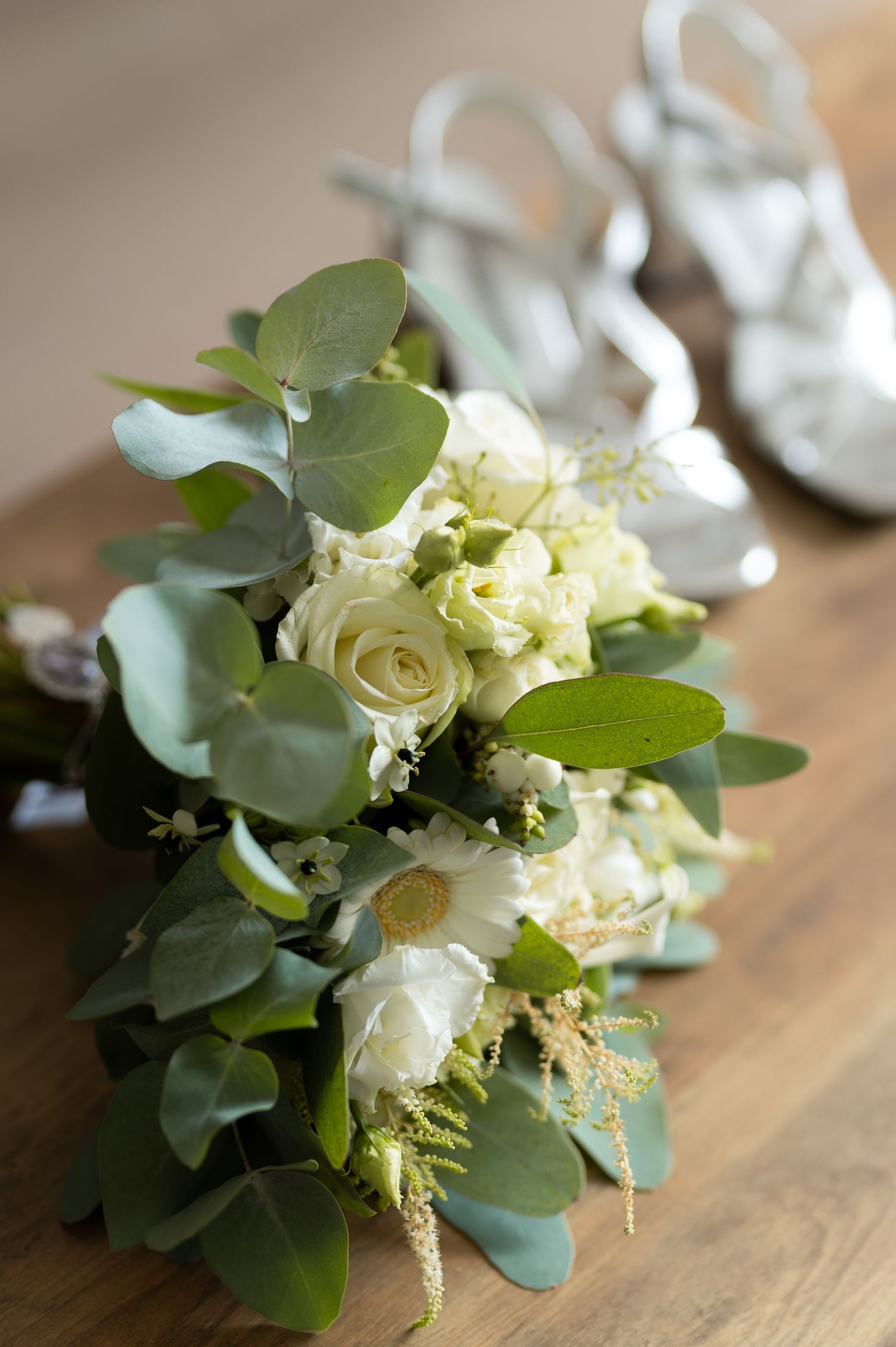 White rose and eucalyptus wedding bouquet on wooden surface with silver bridal heels in background.