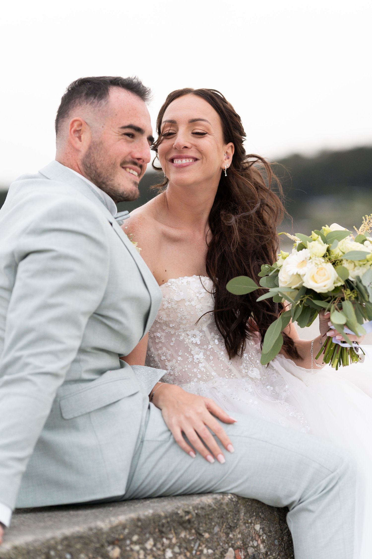 A bride and groom sit closely together outdoors, she holds a white floral bouquet, wearing a lace gown as he smiles in a light gray suit.