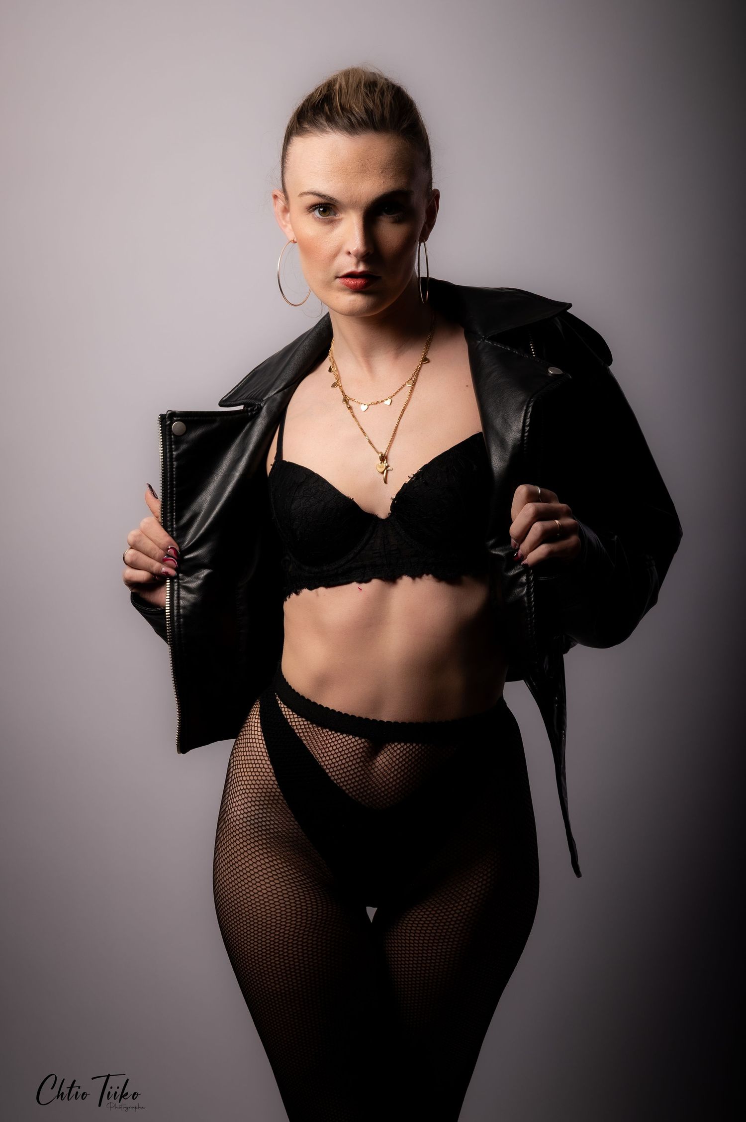 Woman in black lingerie and leather jacket poses confidently wearing gold necklace and hoop earrings in studio.