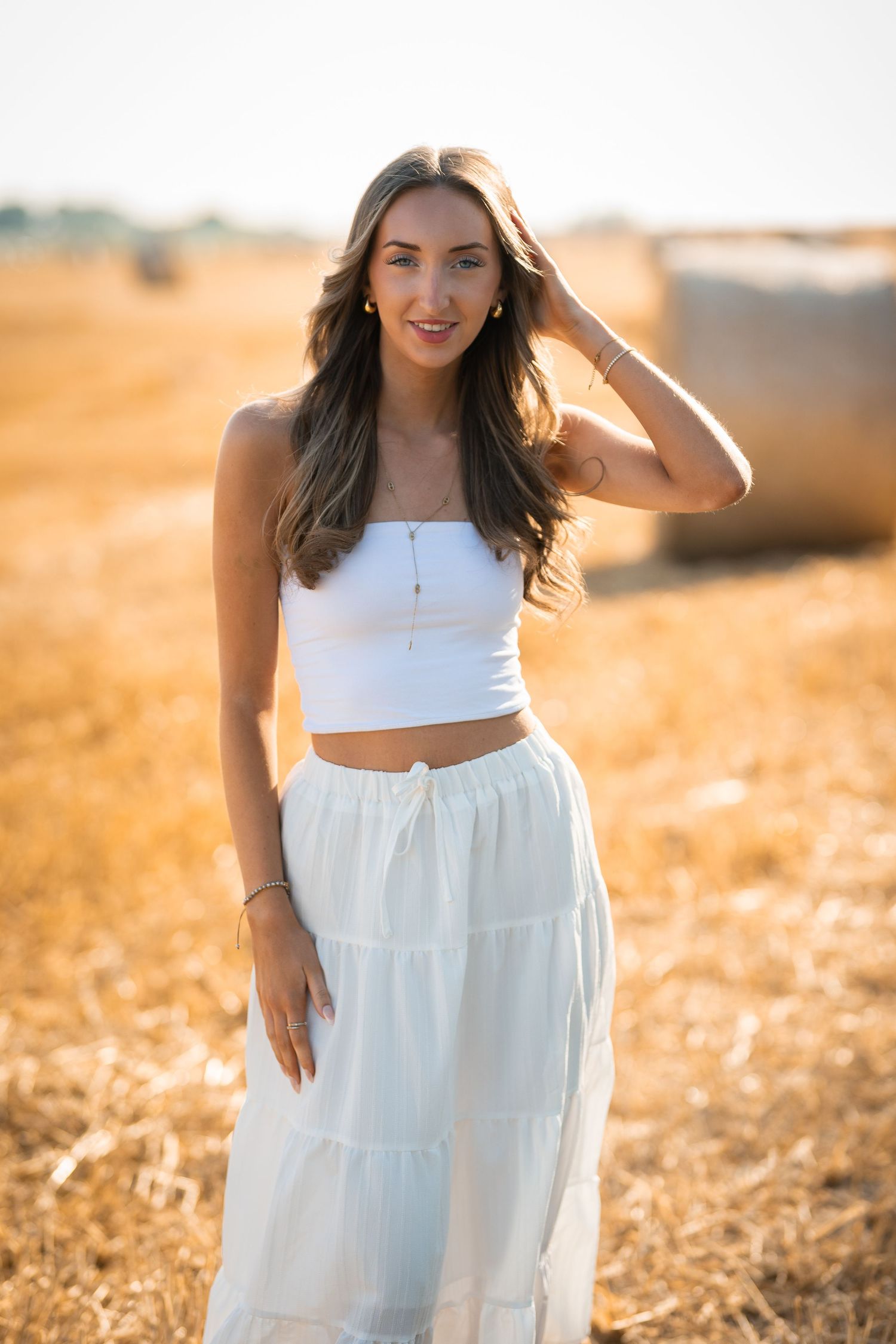 Young woman in white crop top and maxi skirt poses in golden wheat field with hay bales at sunset.