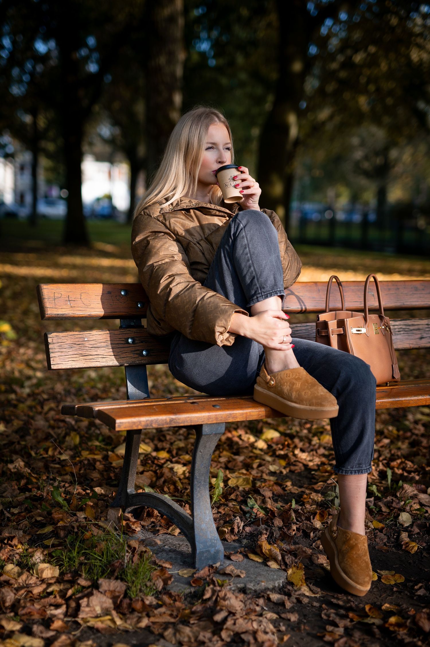 Woman in brown jacket sipping coffee on park bench surrounded by autumn leaves.
