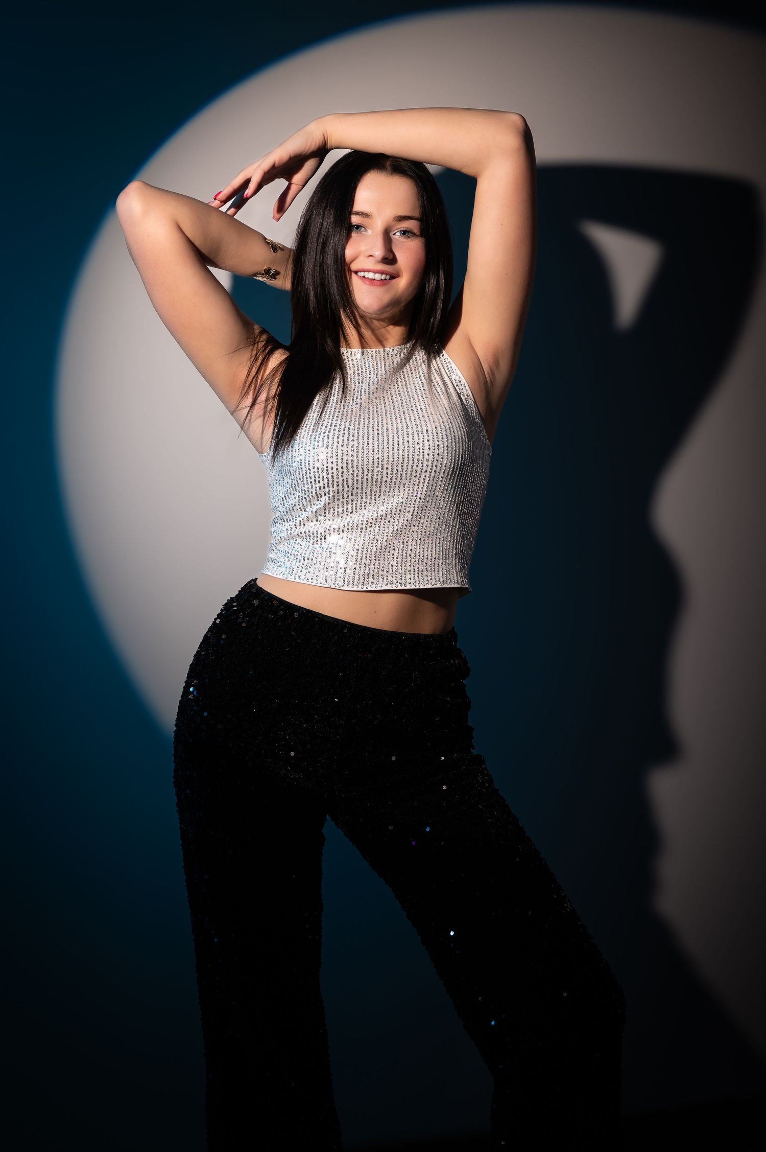 Young woman in silver crop top and black sequin pants poses confidently under spotlight against blue backdrop.