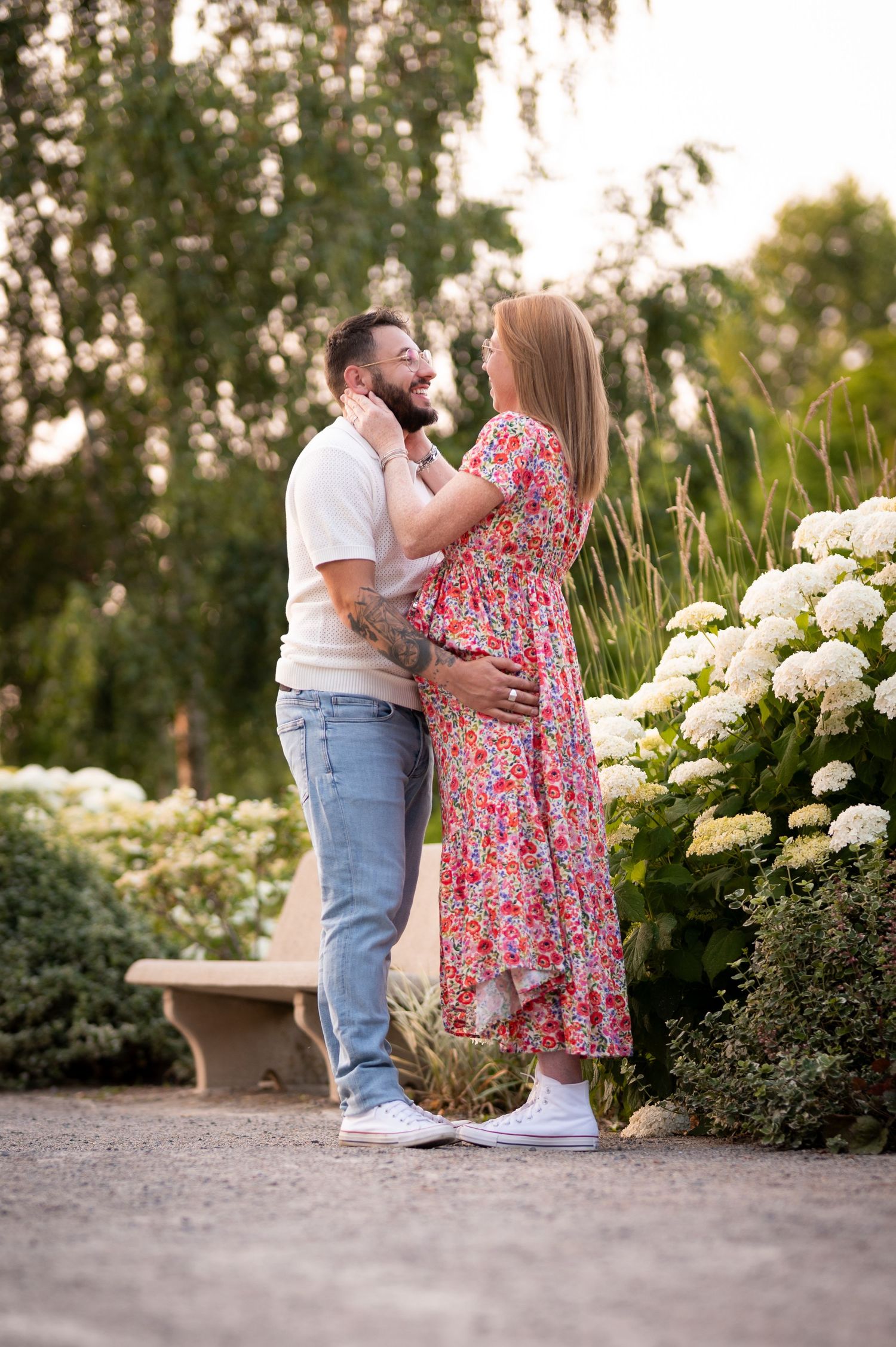 A couple embraces in a lush garden surrounded by white hydrangeas, the woman in a floral dress touching the man's face.