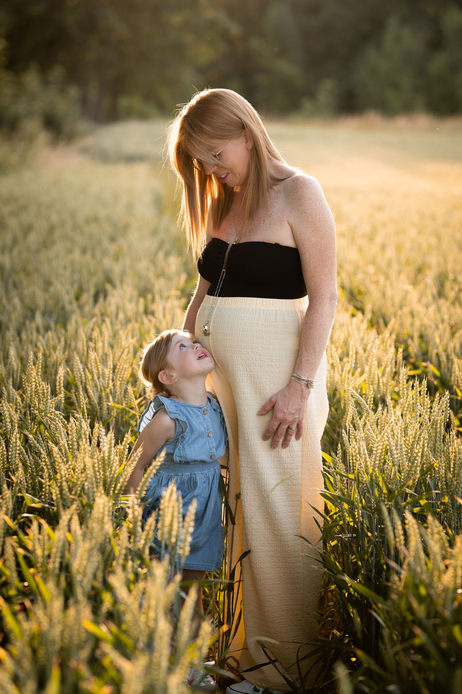 Pregnant woman in a wheat field at golden hour with young girl looking up at her baby bump.