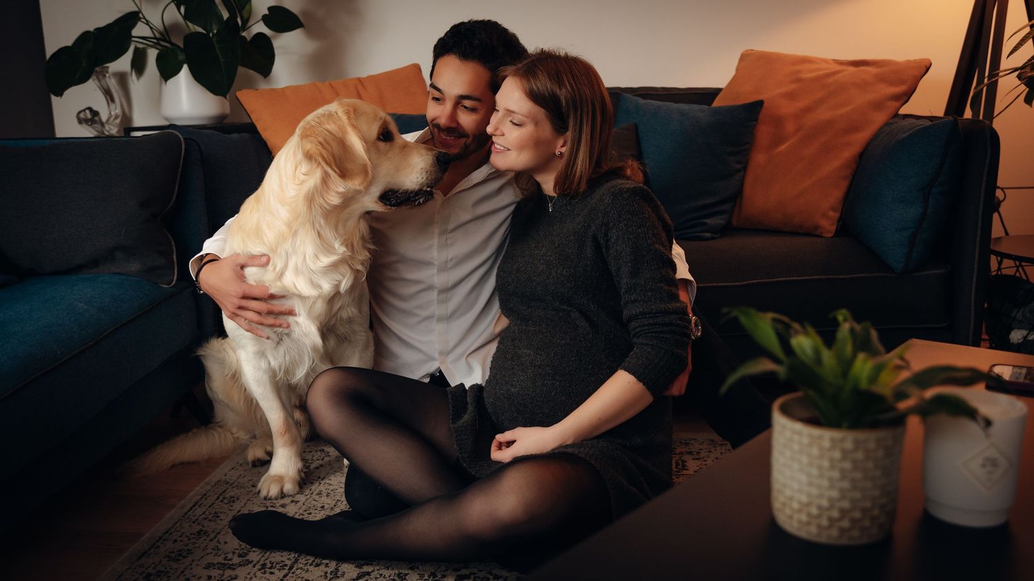Couple sitting on floor with golden retriever dog in cozy living room with blue sofa and warm lighting.