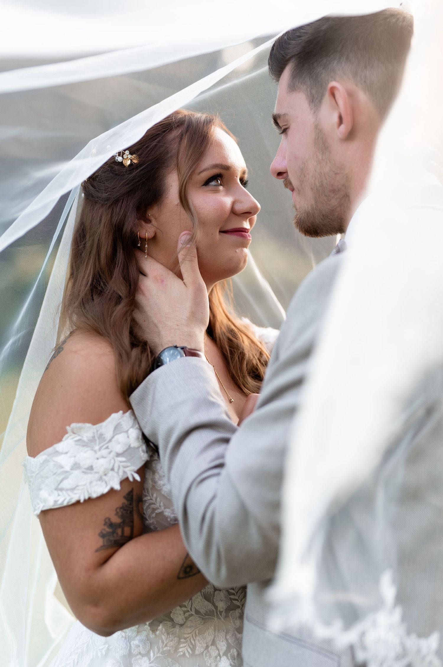 Groom tenderly touching bride's face under flowing veil as she gazes up at him lovingly outdoors.
