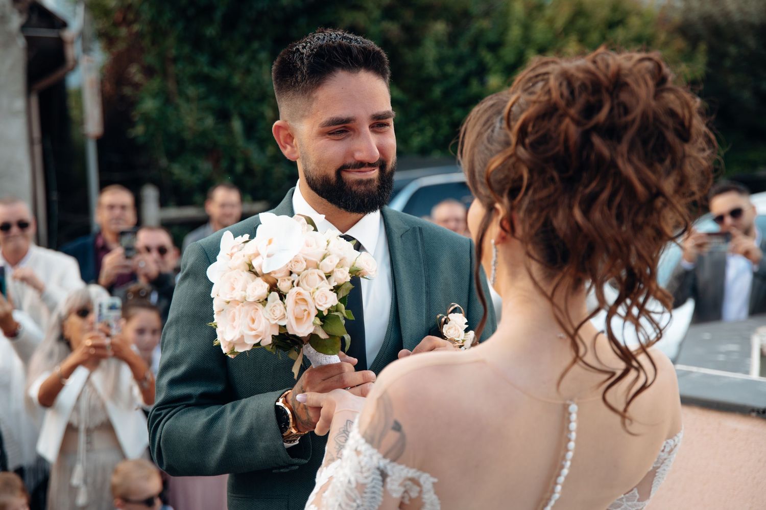 Smiling groom in teal suit presents white rose bouquet to bride during outdoor wedding ceremony.