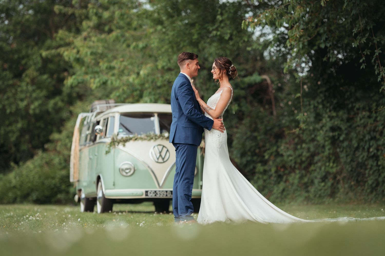 Bride and groom embracing in front of a vintage mint green VW camper van decorated with floral wreath at outdoor wedding.