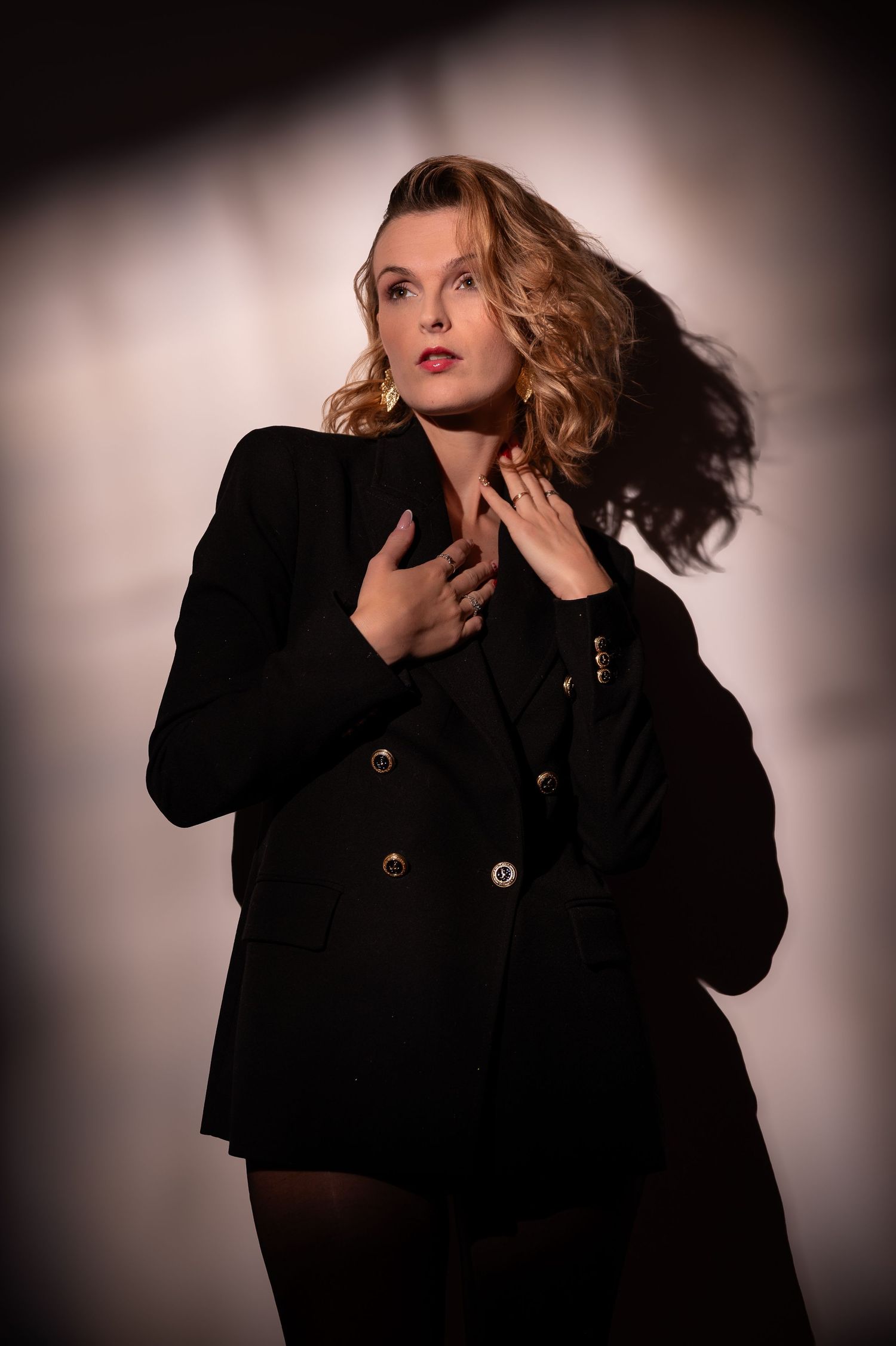 A blonde woman in a black double-breasted blazer poses with flowing hair in dramatic studio lighting.