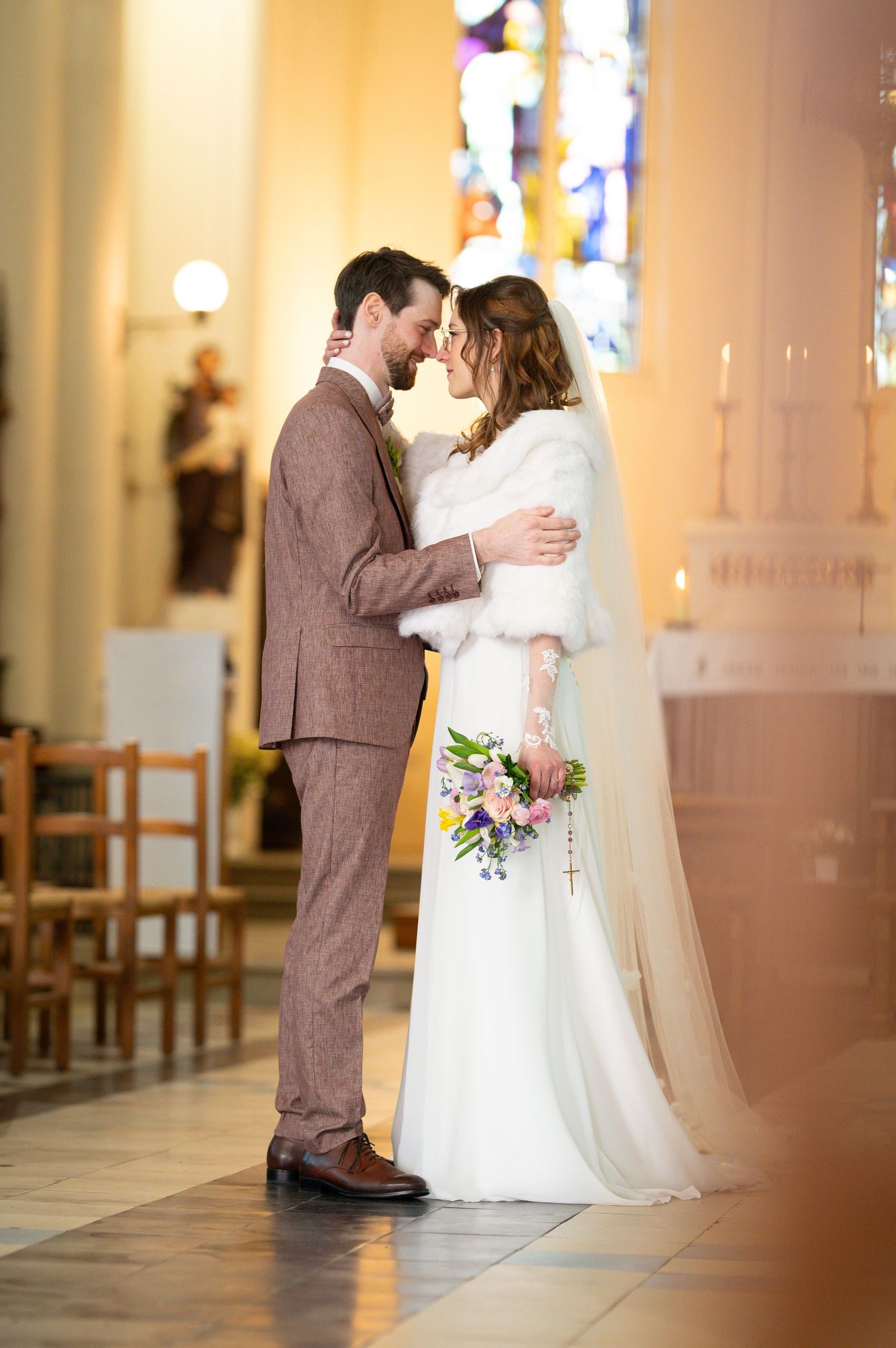 Bride and groom share a romantic moment in a church, bride wearing white gown with fur stole holding colorful bouquet.