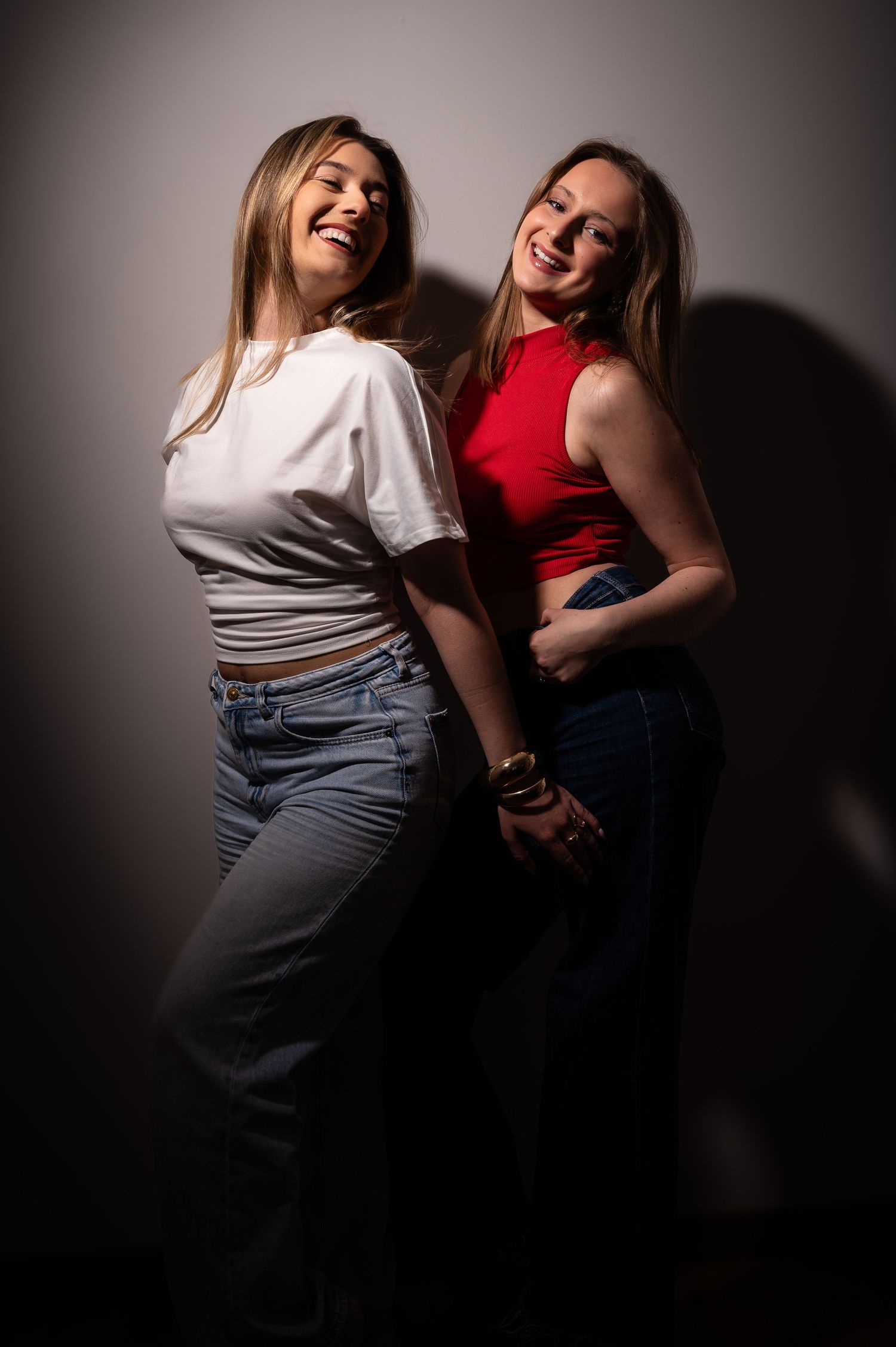 Two smiling young women posing together, one in white tee and one in red crop top, both wearing jeans against a gray backdrop.