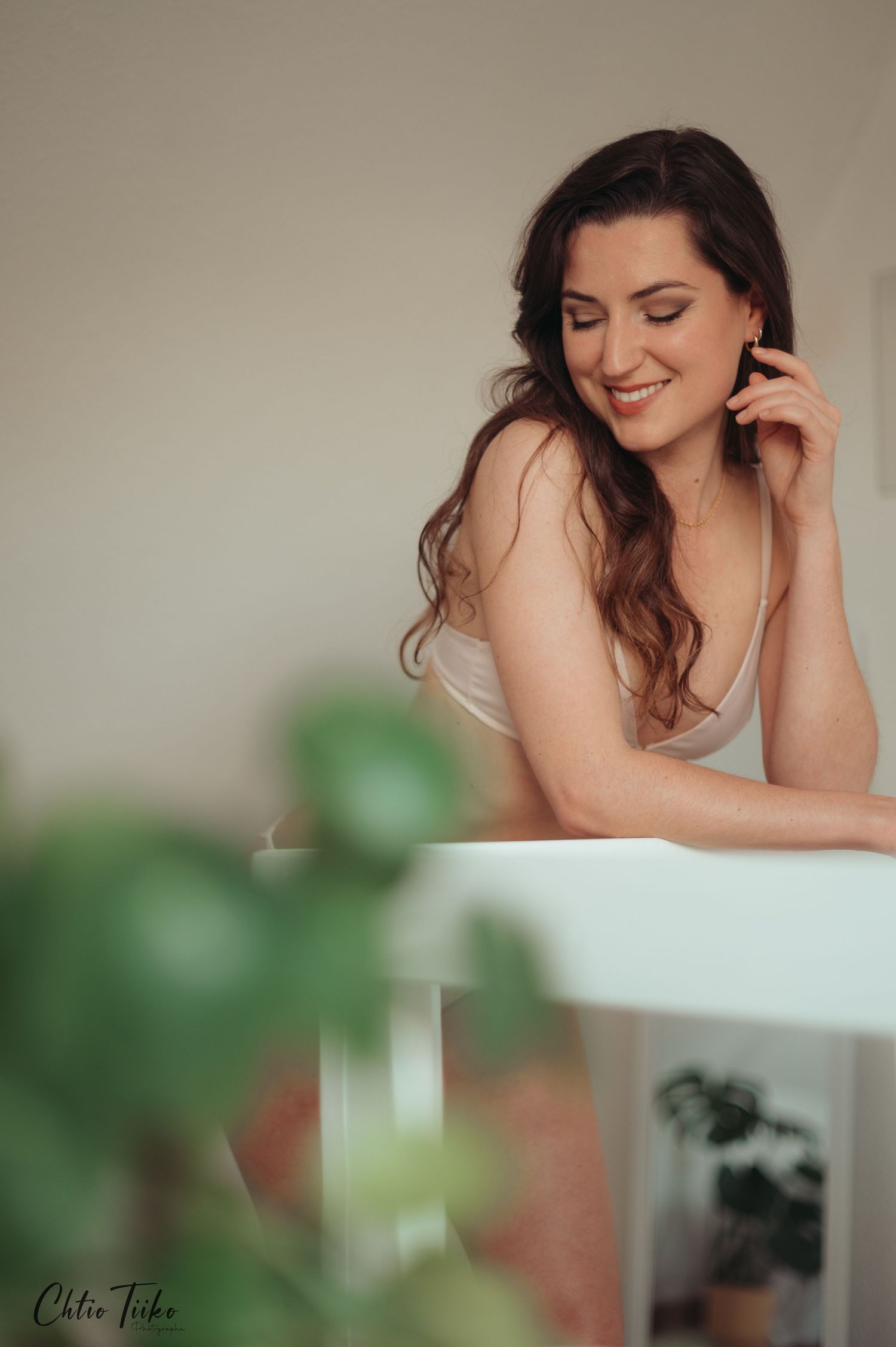Smiling brunette woman in white top leaning on a white table, touching her earring in a bright room.