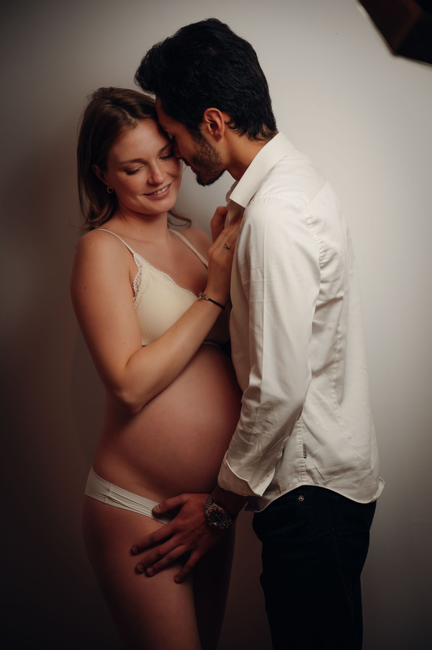 Couple sharing tender moment during maternity photoshoot, pregnant woman in nude lingerie embracing man in white shirt.