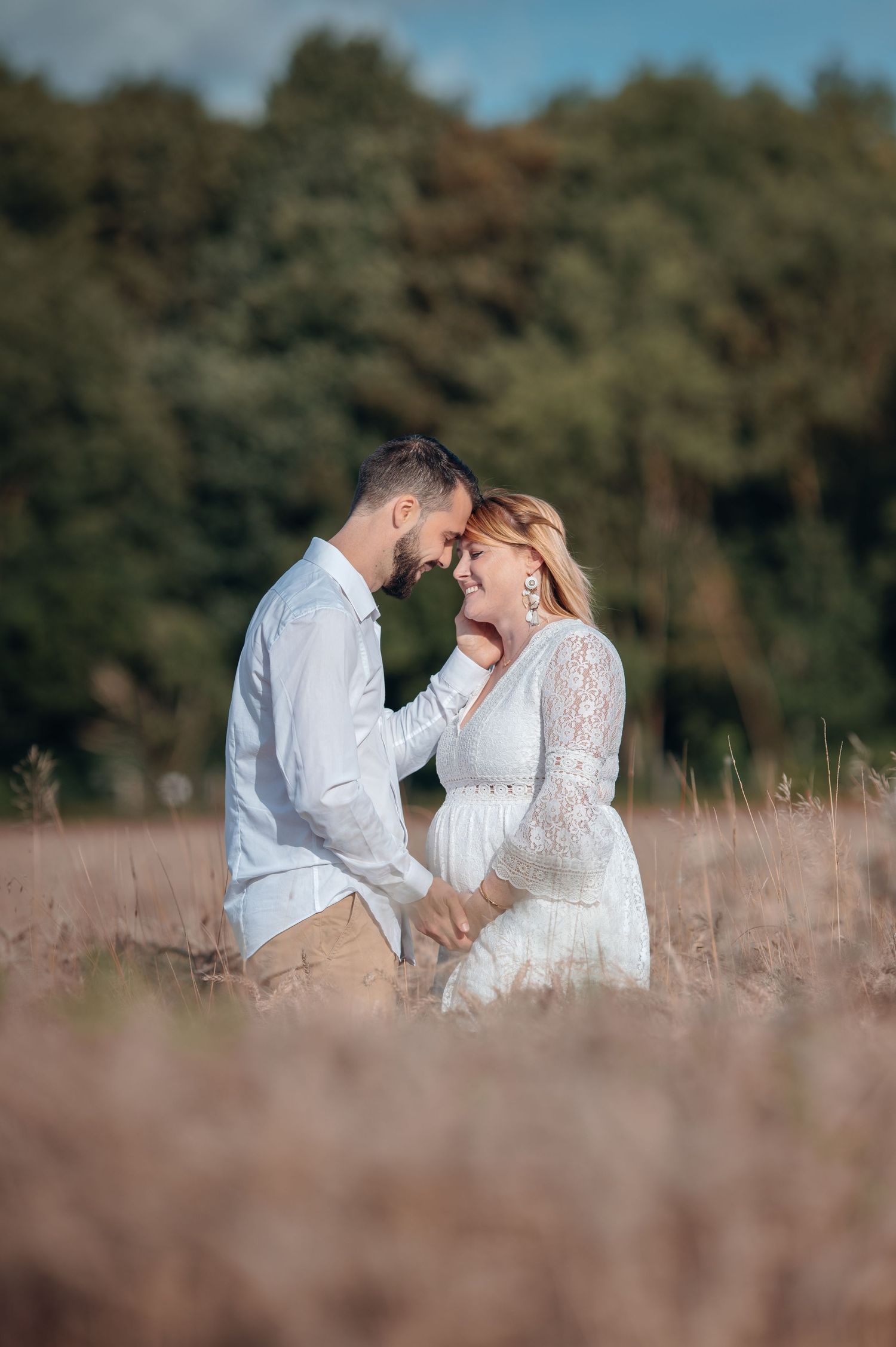 A couple shares a tender moment in a golden field, the woman in a white lace dress, with trees in the background.