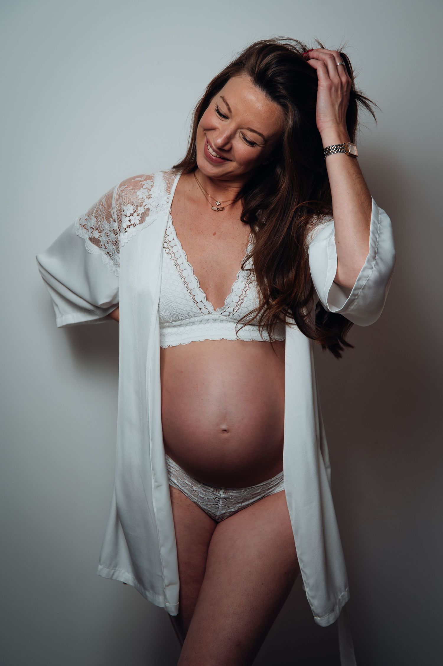 Smiling pregnant woman in white lace lingerie and open robe, posing with hand in hair against light background.