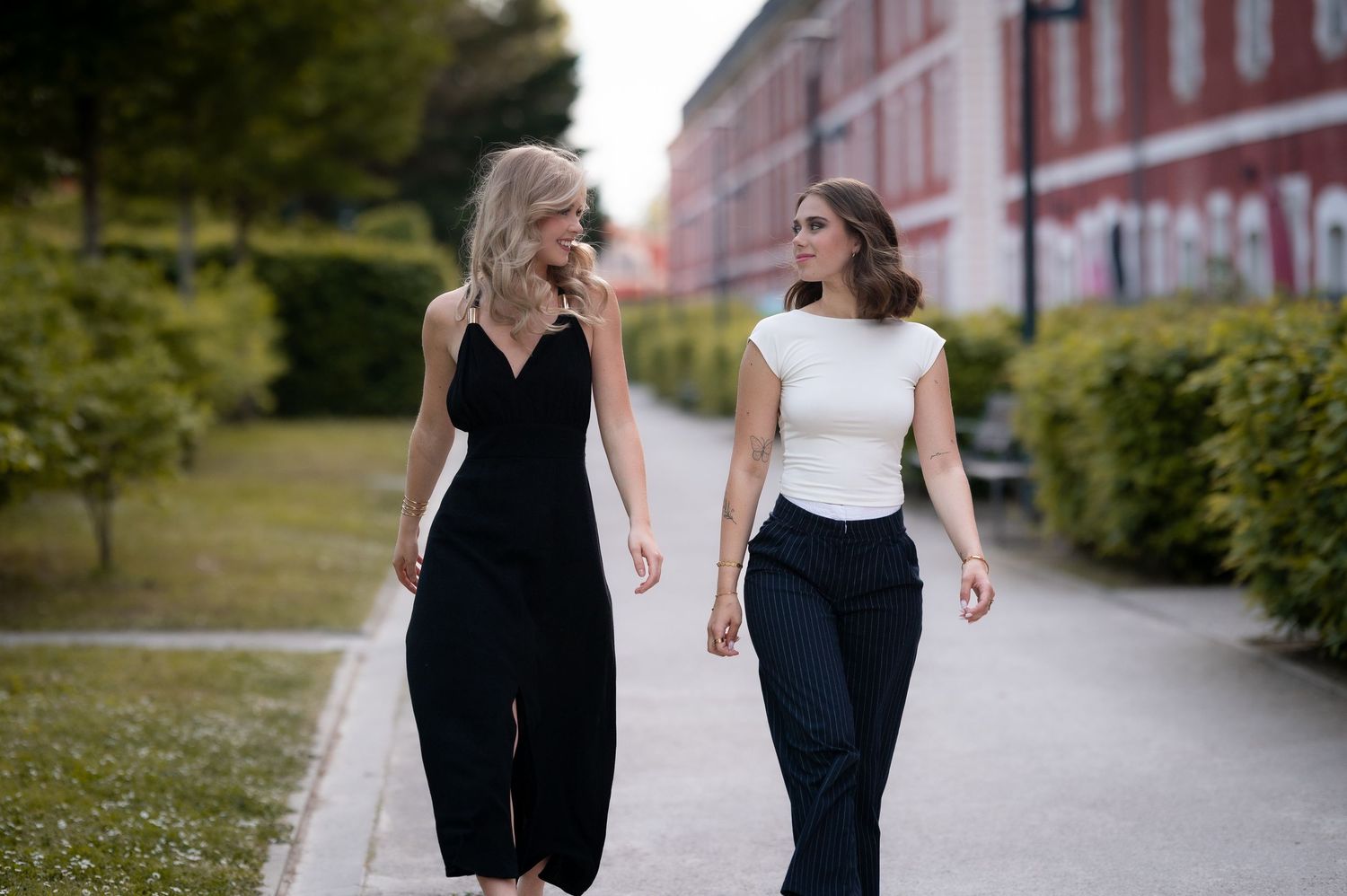 Two elegant women walking outdoors on a path, one in black jumpsuit and one in white top with black pants.