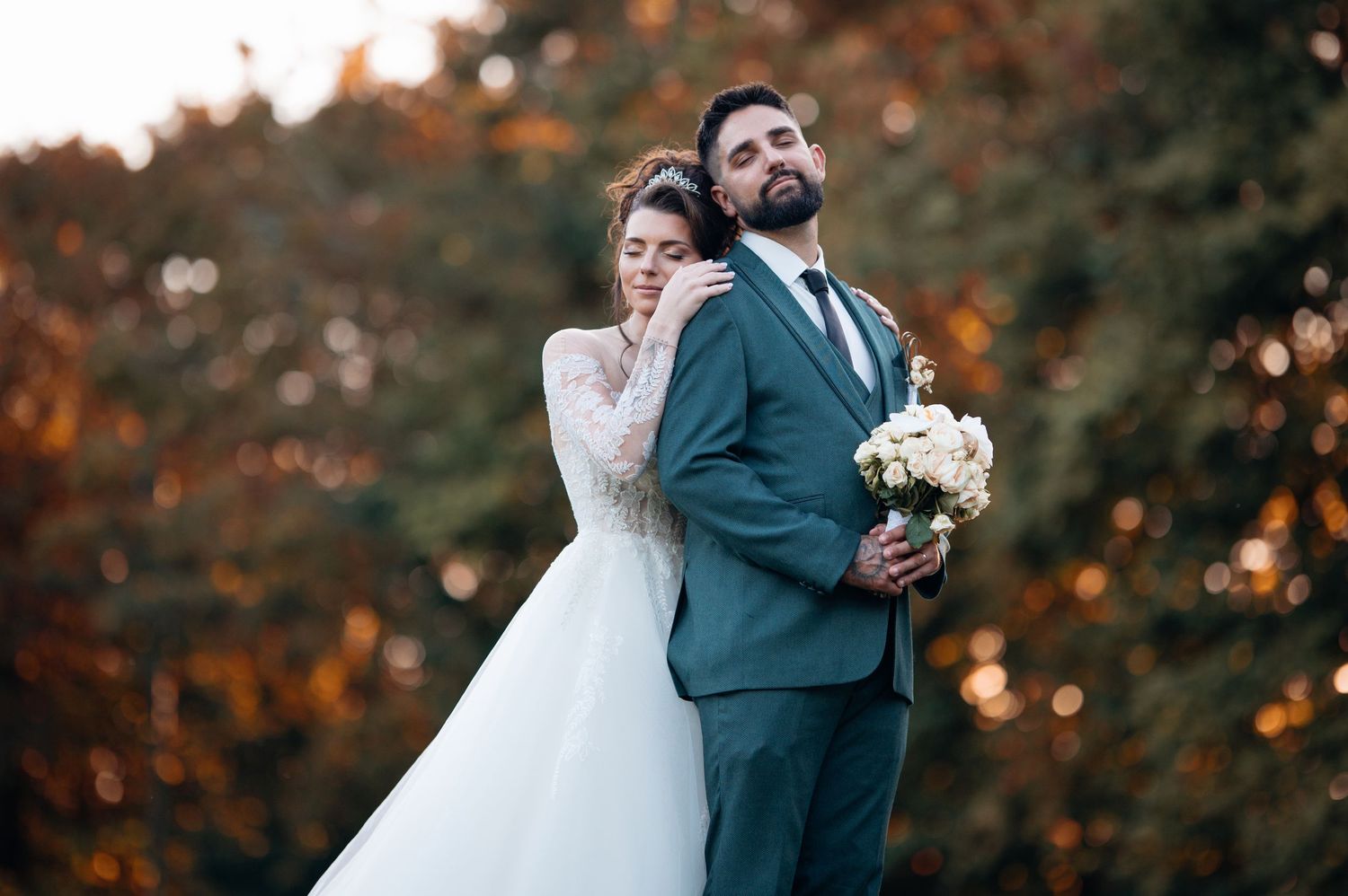 Bride in lace gown embraces groom in teal suit holding white bouquet against colorful autumn foliage.