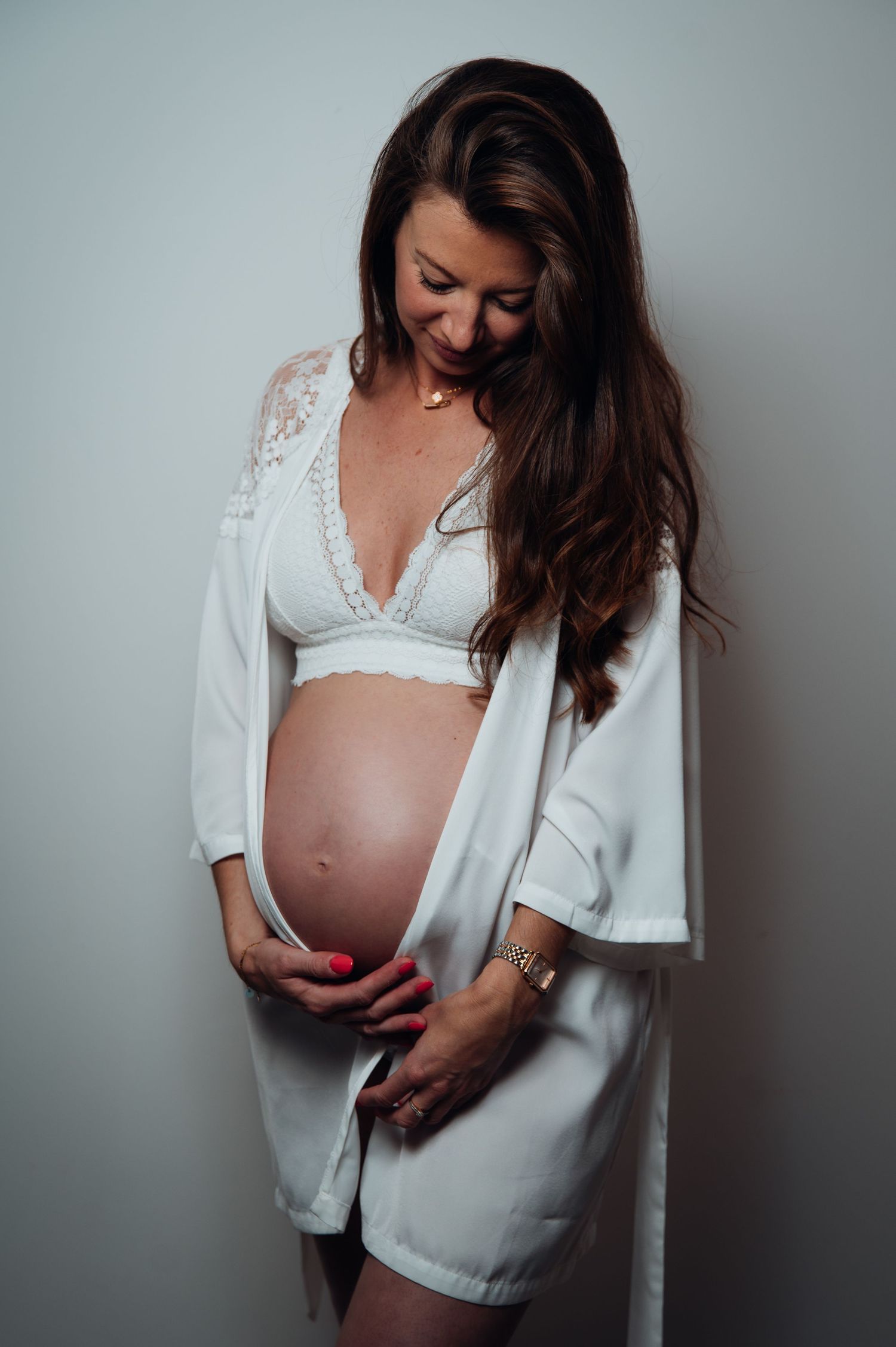 Pregnant woman in white lace bralette and robe cradling her baby bump, smiling downward in a studio setting.