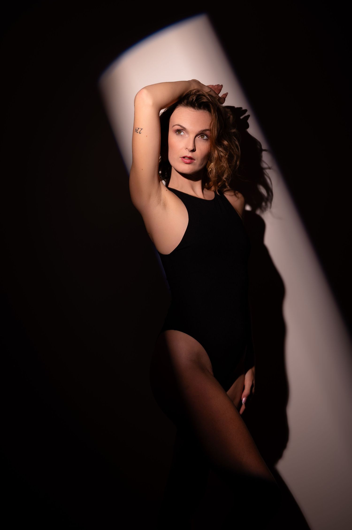 Woman in black bodysuit posing dramatically in a narrow beam of light against a dark studio background.