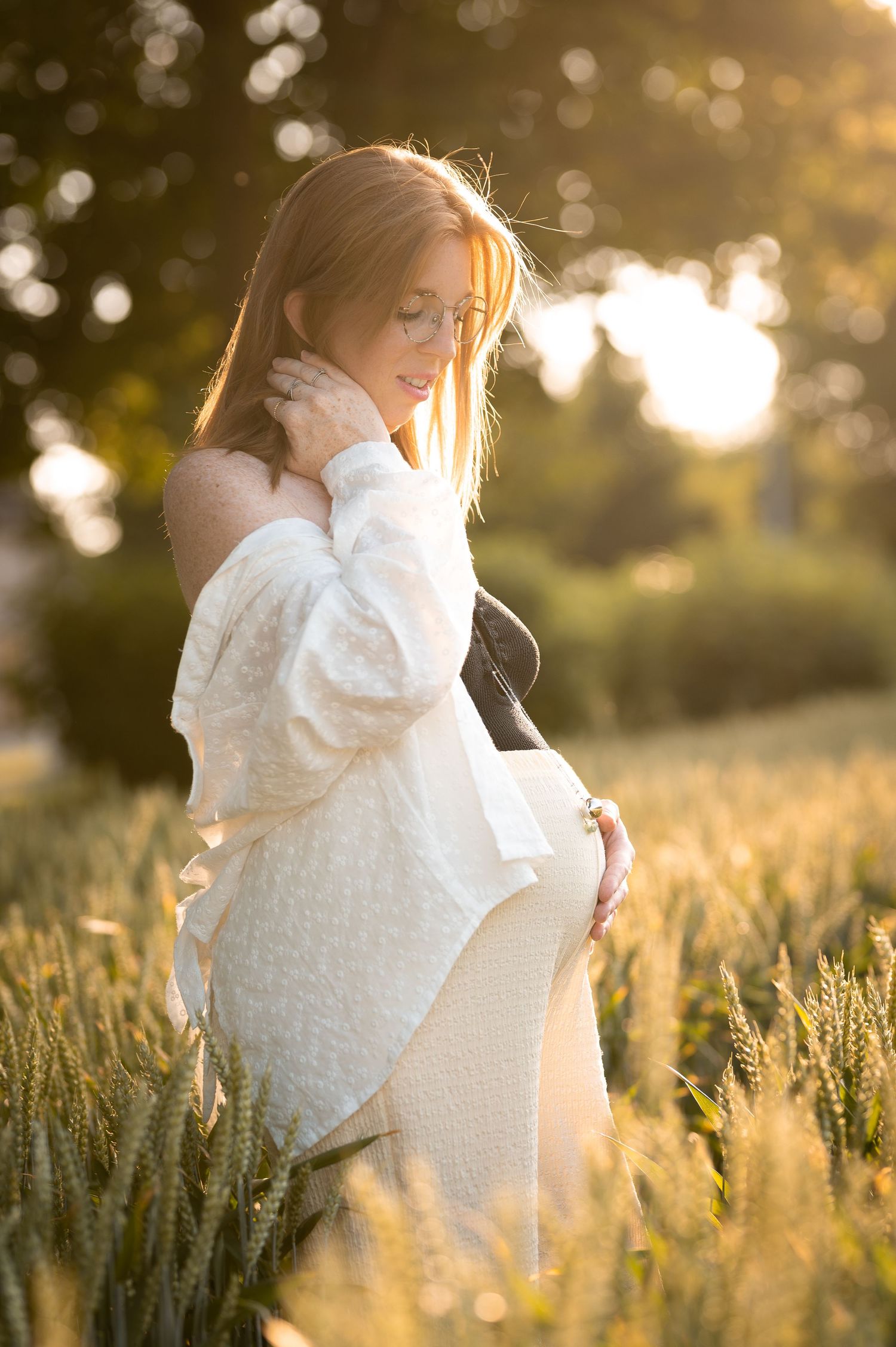 Pregnant woman in white dress standing in golden wheat field at sunset, cradling her baby bump with glowing backlight.