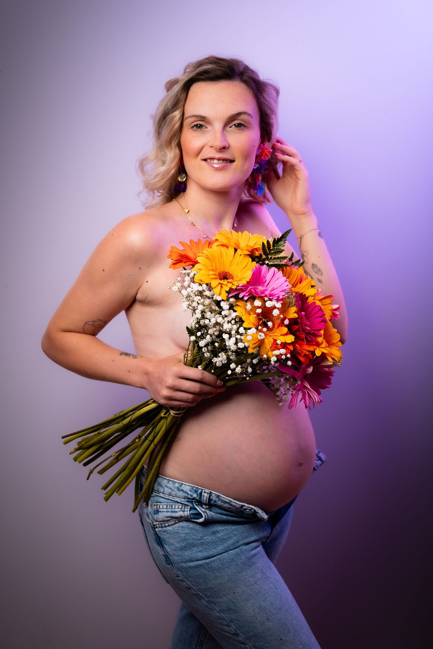 Smiling pregnant woman holding colorful gerbera flower bouquet, posing topless in jeans against purple studio background.