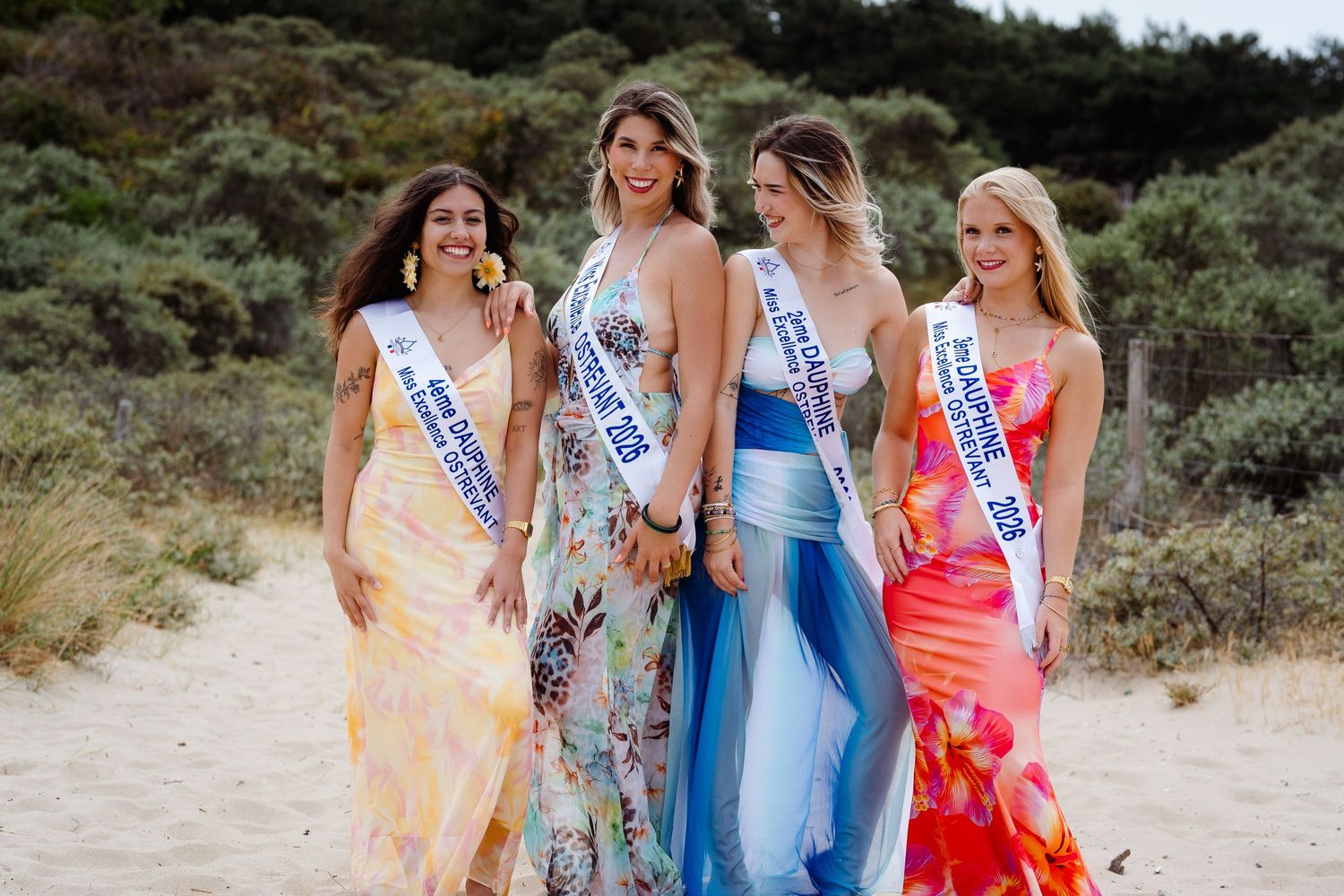 Four women in elegant gowns wearing pageant sashes pose together on a sandy beach with greenery in the background.