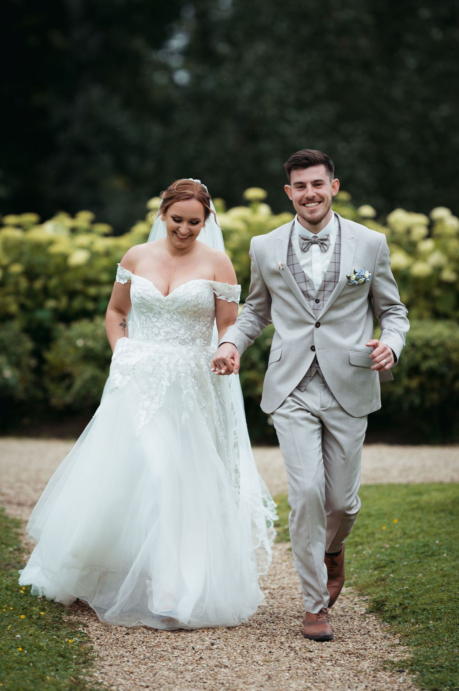 Bride in off-shoulder lace gown and groom in grey suit holding hands walking on garden path at wedding.