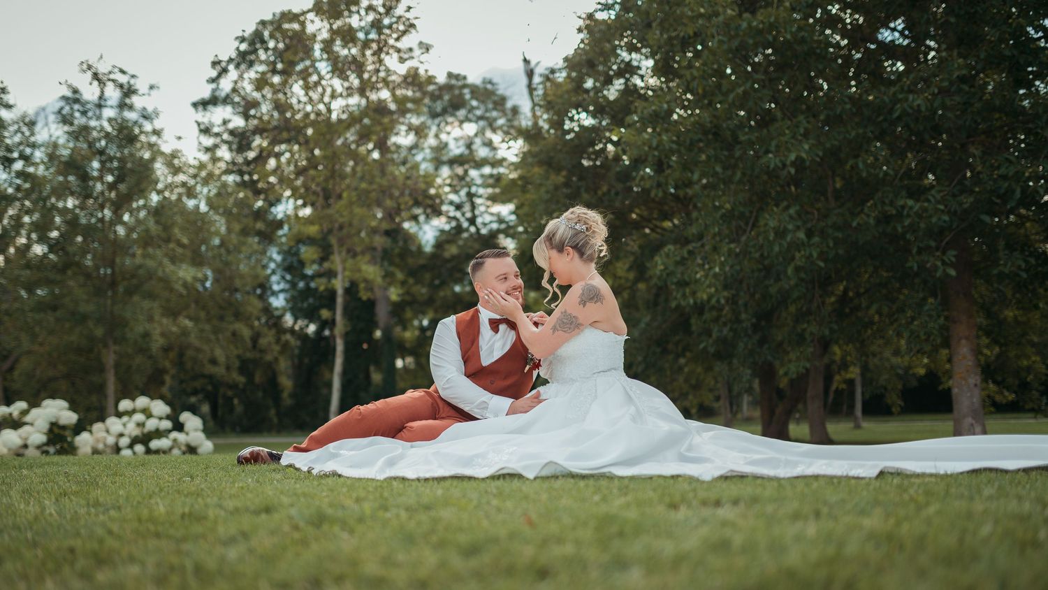 A bride and groom sit together on a white blanket in a lush green park, sharing a romantic moment on their wedding day.